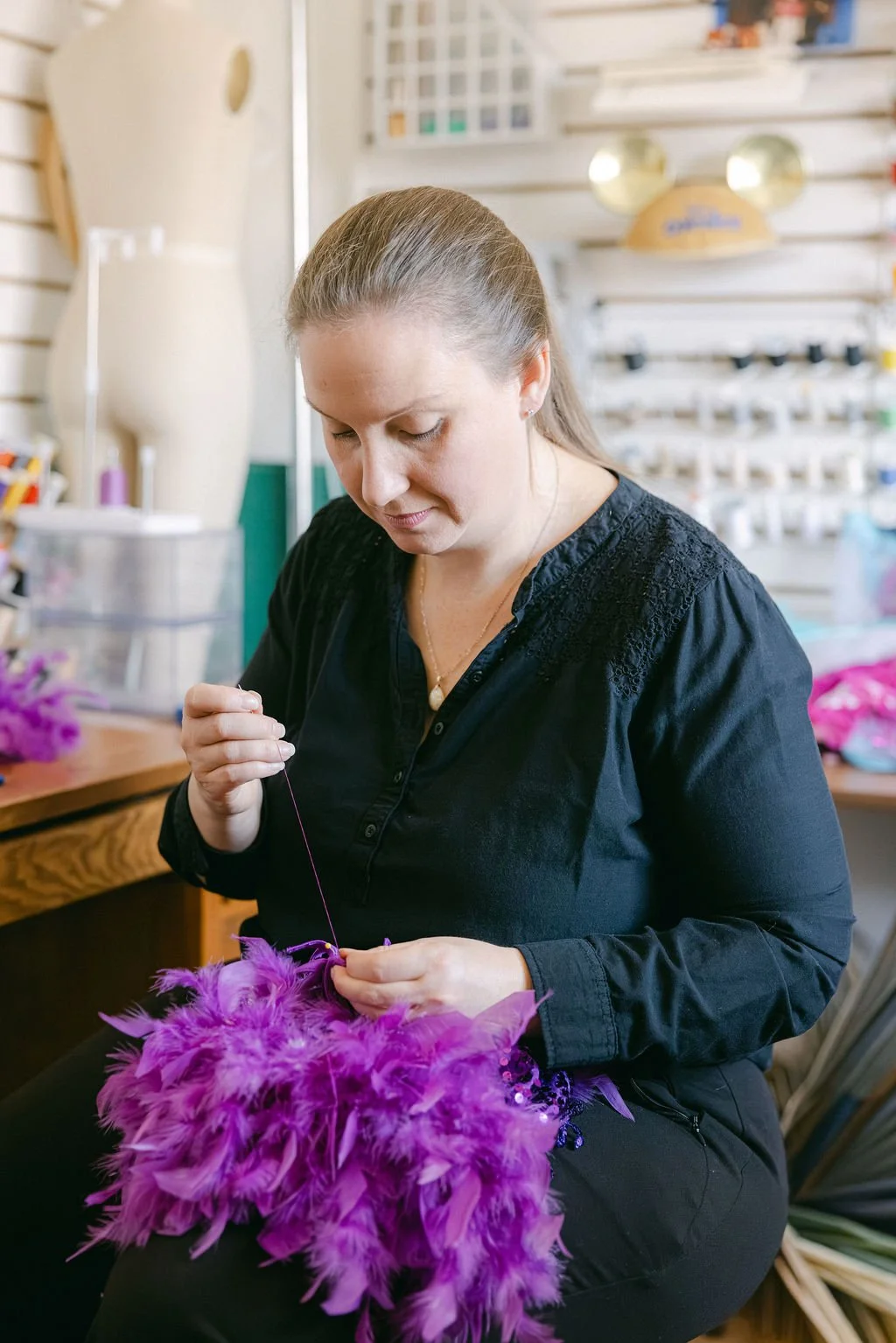 A woman working on a purple feathered craft at a workspace with crafting supplies and tools.