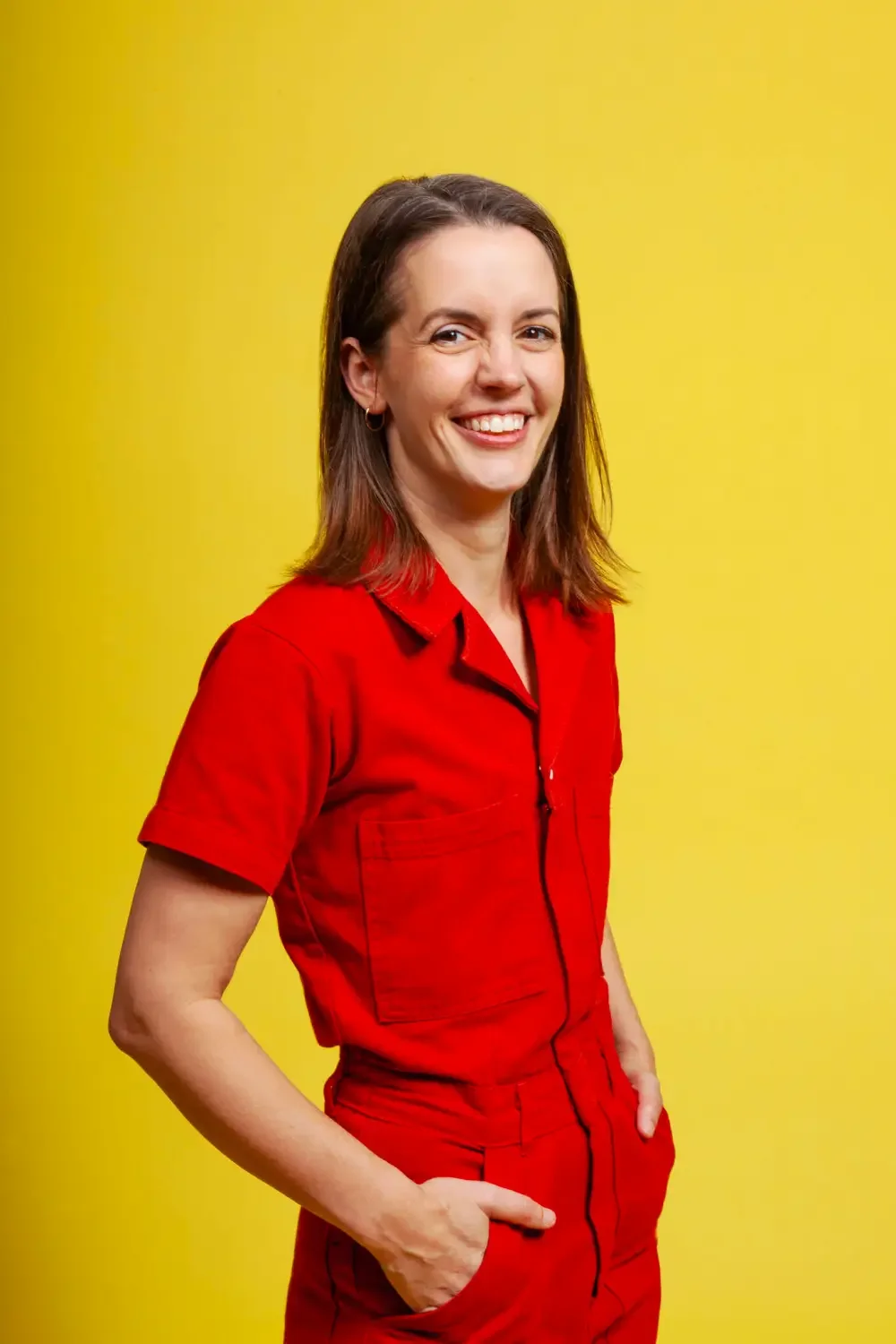 A woman with shoulder-length brown hair, wearing a red shirt and pants, smiling with hands in pockets, standing against a yellow background.