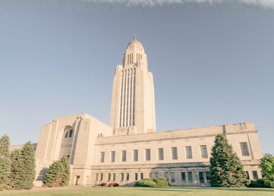 The Nebraska State Capitol building with a tall central tower, surrounded by trees and a clear sky.
