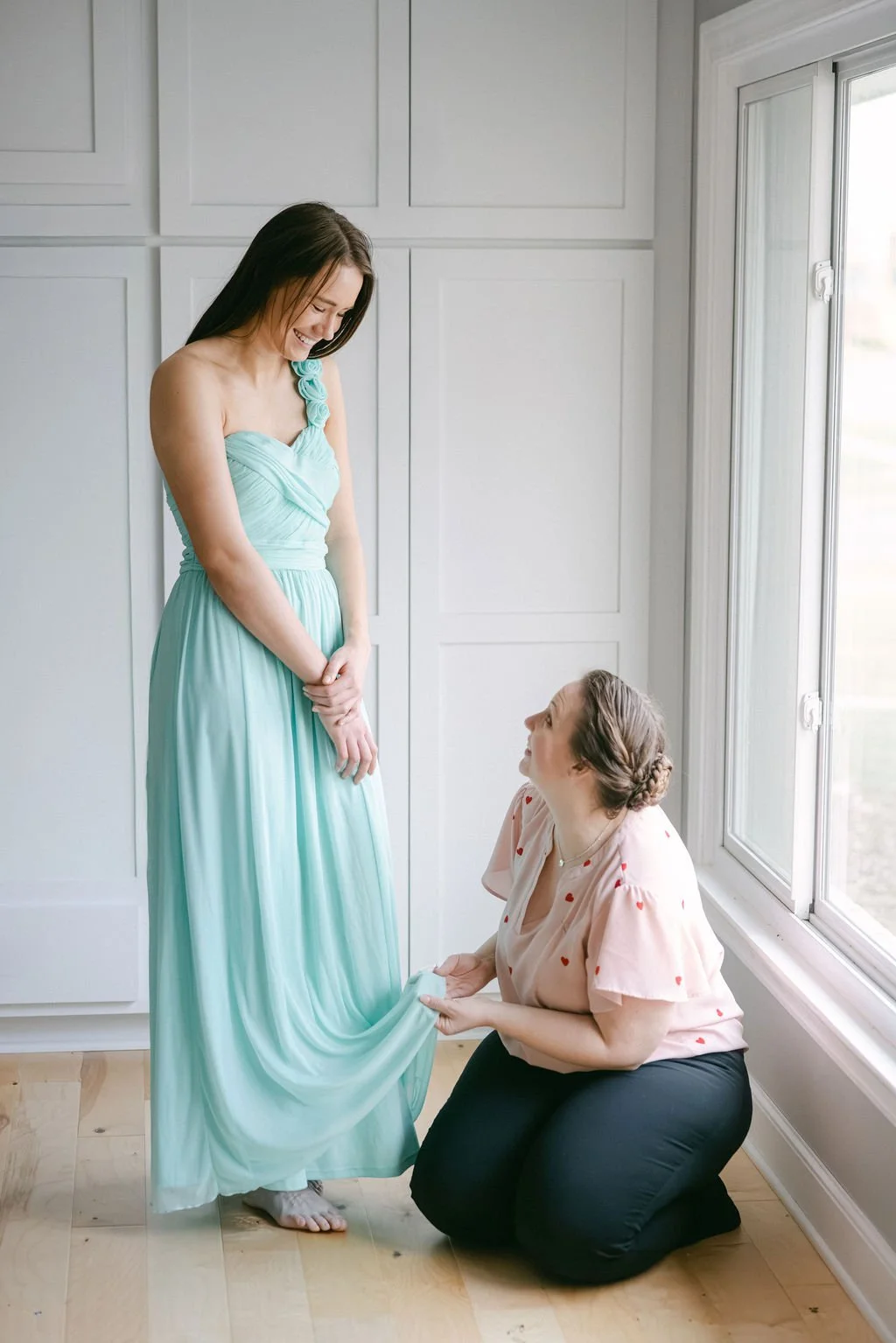 A woman in a light blue dress smiles as she is helped with her dress by another woman who is kneeling on the wooden floor, wearing a pink blouse with red hearts and black pants. They are in a bright room with a large window.