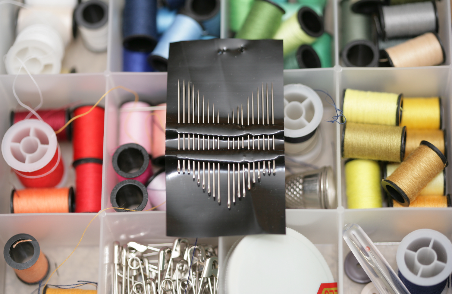 A sewing kit containing spools of thread in various colors, sewing pins on a black pin holder, and other sewing accessories organized in a plastic compartment.