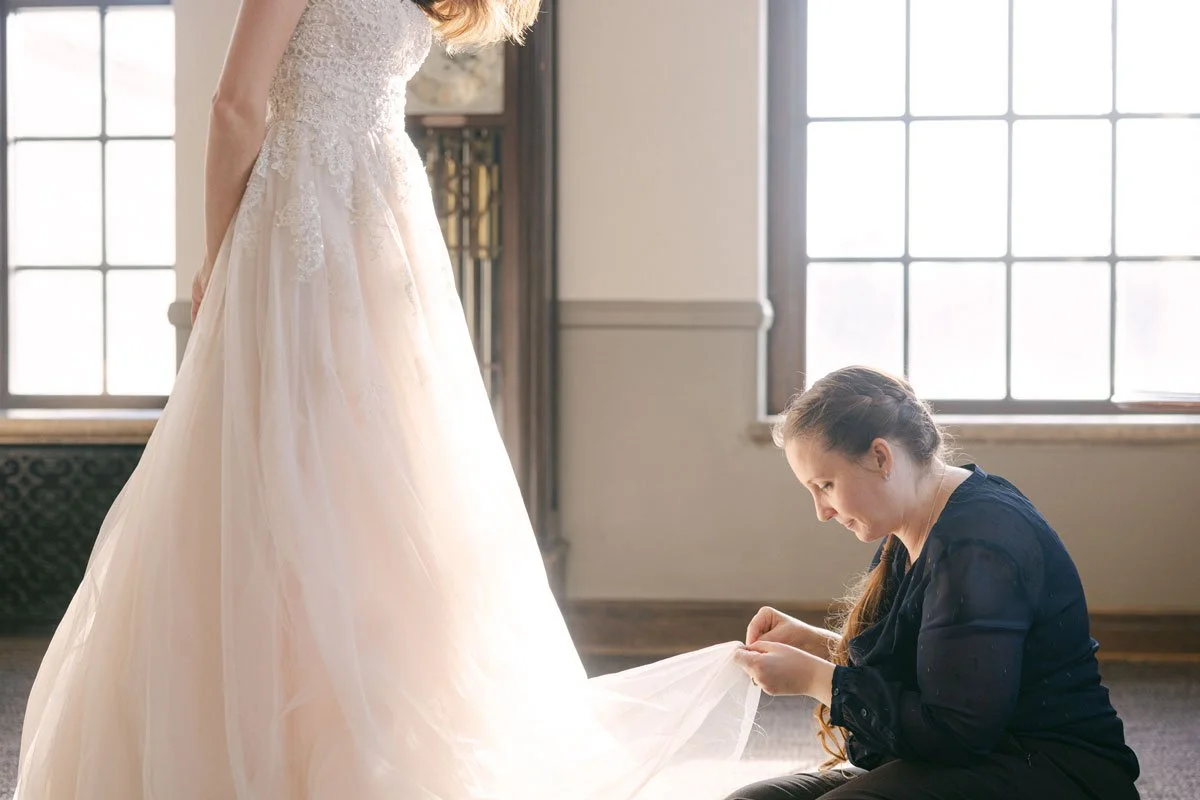 A woman in black sits on the floor, adjusting the train of a wedding gown worn by another woman, inside a room with large windows.