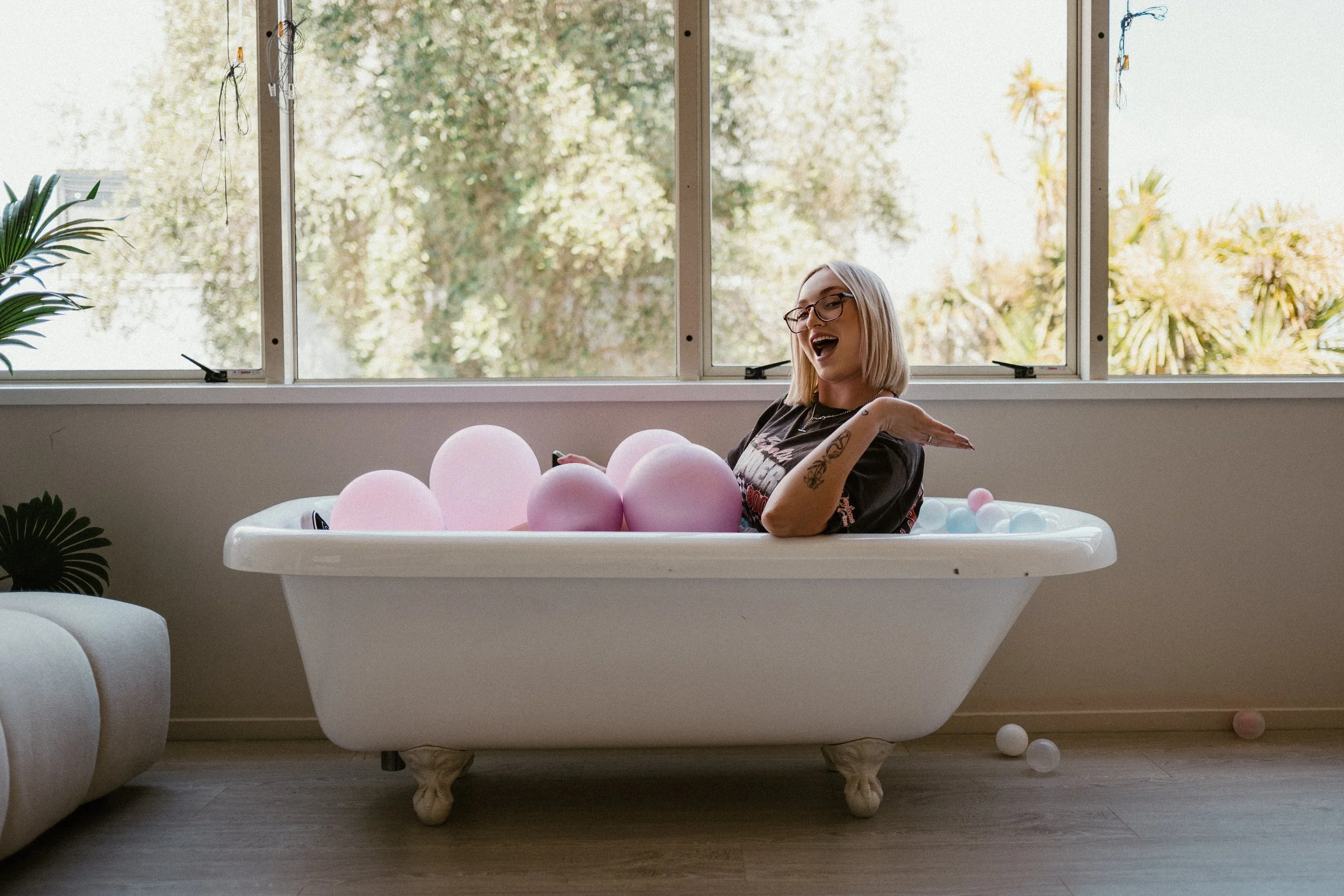 A woman enjoying herself in a vintage bathtub filled with pink and light blue balls, sitting in front of a large window with a view of trees outside.