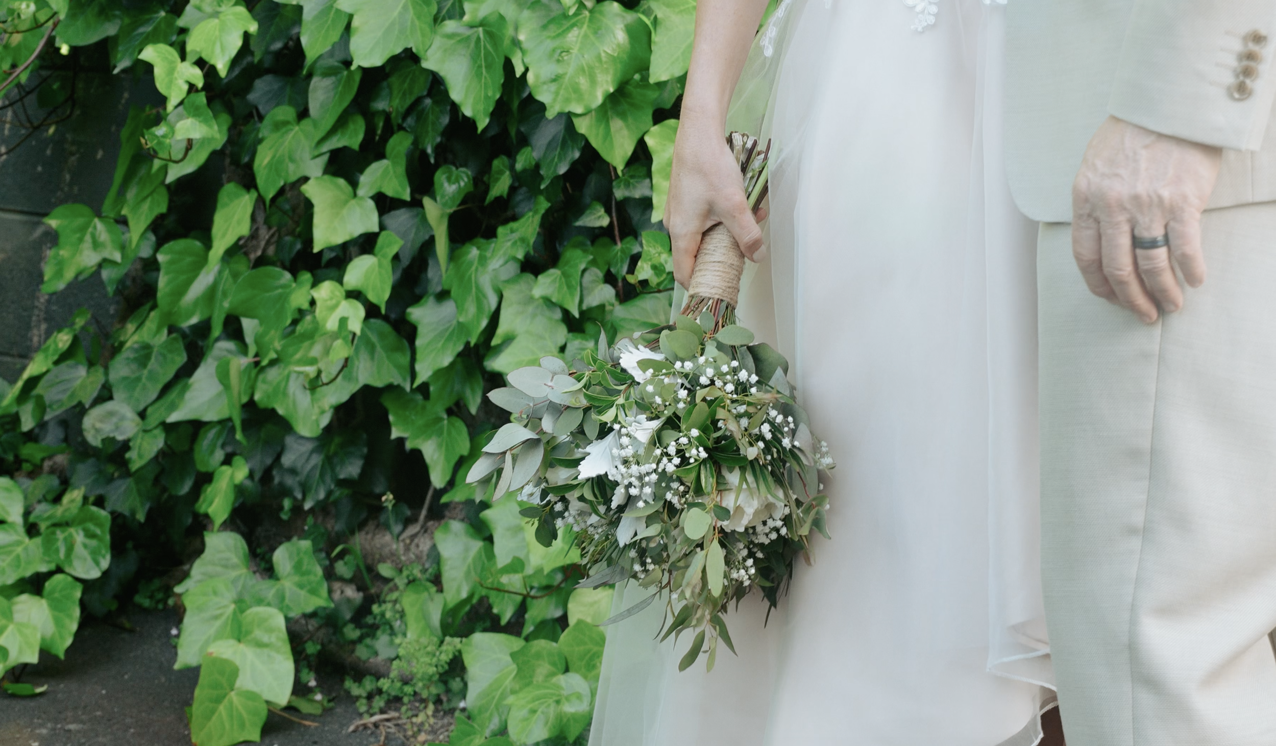 Special events. A bride holding a bouquet of white and green flowers, standing next to a groom in a light-colored suit, with a lush green leafy background.