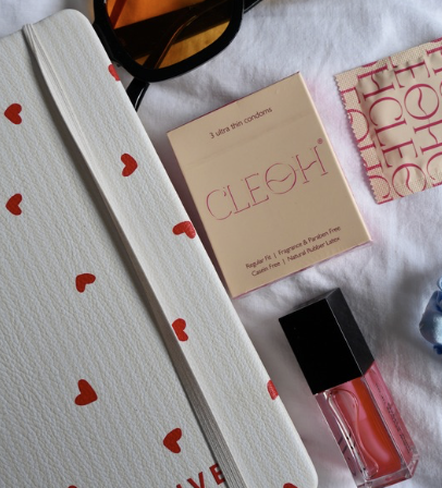 Flat lay of personal items including a white textured notebook with red heart designs, a small pink box labeled 'CLEOH' with condom brand information, a pink and white patterned cloth, a black sunglasses case, pink lipstick, and a pair of blue underwear, all arranged on a white fabric surface.