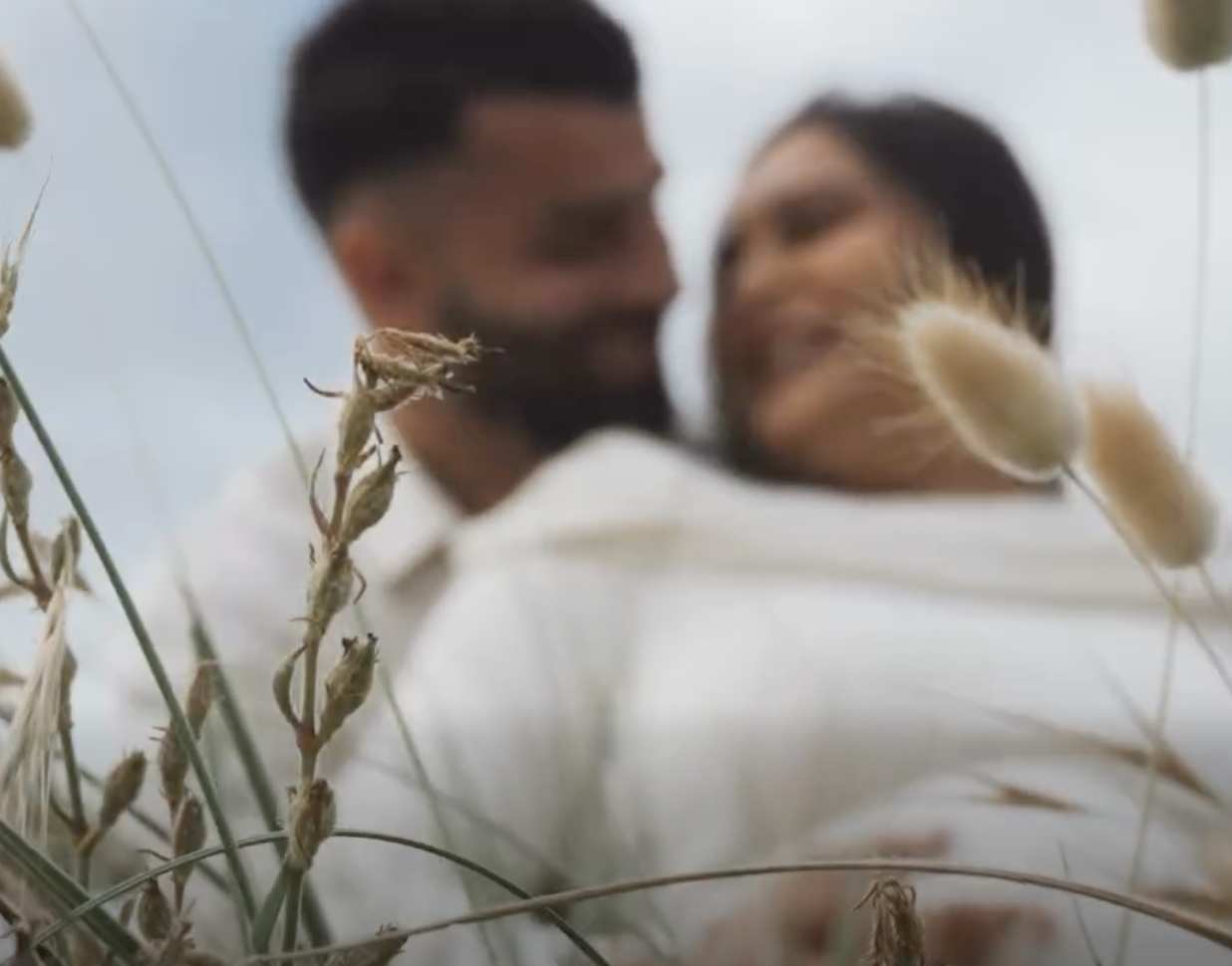 A blurred picture of a couple embracing in a field of tall dry grass, with some dried grass in the foreground.