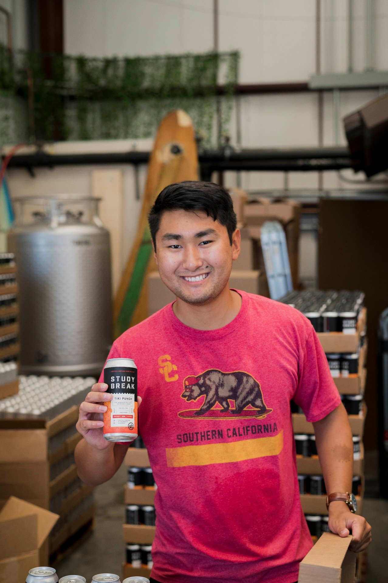 A young man wearing a red t-shirt with a bear and the words "Southern California" is smiling and holding a can with a label that says "Study Break" and "Tiki Punch". He is in a warehouse or storage area surrounded by boxes and bottles.