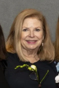 A smiling middle-aged woman with blonde hair, wearing a dark top with a small green plant pinned to it, standing against a gray background.