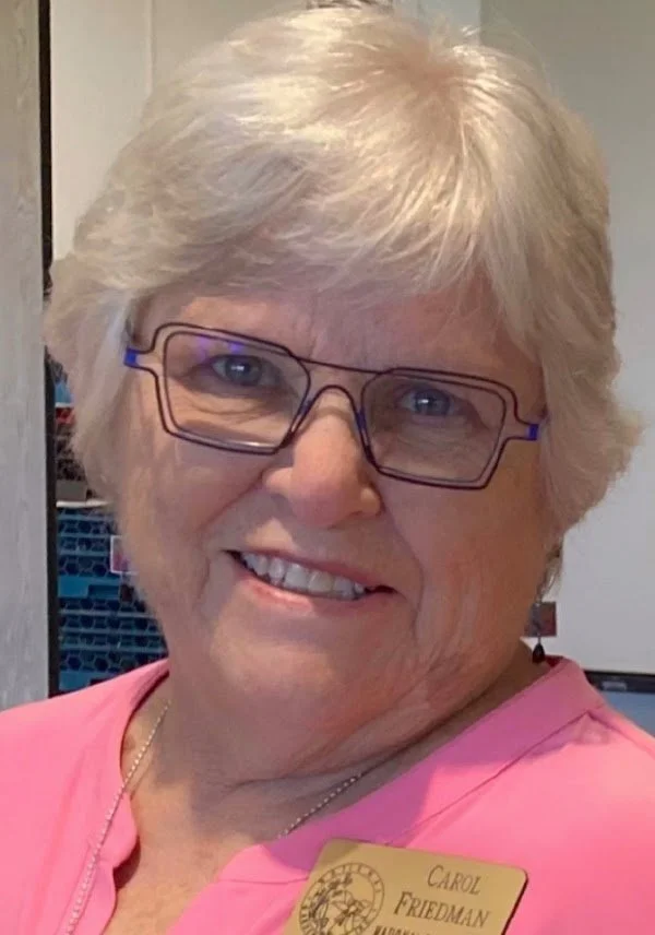 A smiling older woman with short, white hair and glasses, wearing a pink shirt and a name tag that reads 'Carol Friedman'.