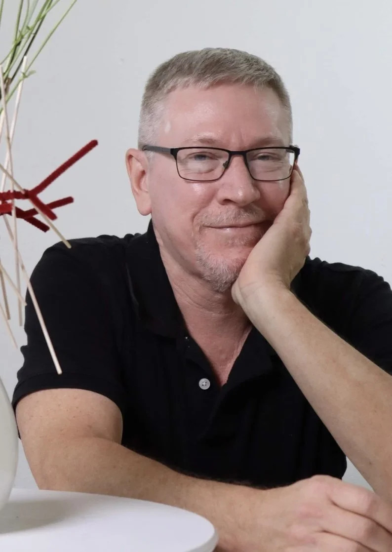 A middle-aged man with short, gray hair and glasses, wearing a black shirt, sitting at a table with a light-colored background, resting his face on his hand and smiling.