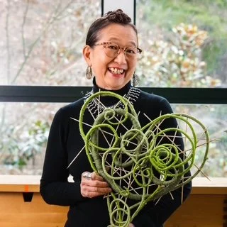 Woman with glasses and earrings smiling while holding a green spiraled sculpture in a room with a large window and greenery outside.