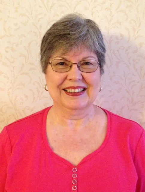 Smiling elderly woman with short gray hair, wearing glasses, earrings, and a pink top, standing in front of a beige wallpapered wall.