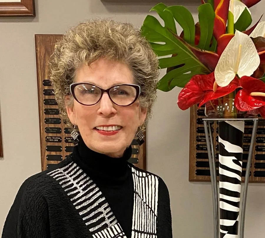 Women with short curly gray hair and glasses smiling, wearing a black and white striped top, standing next to a large floral arrangement with red and white flowers in a tall vase.