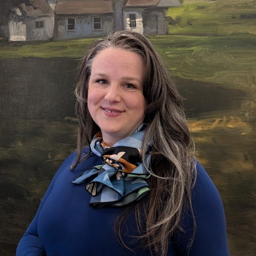 A woman with long, wavy brown hair and light skin, smiling, wearing a dark blue top and a multicolored scarf, standing in front of a painted landscape background depicting houses and greenery.