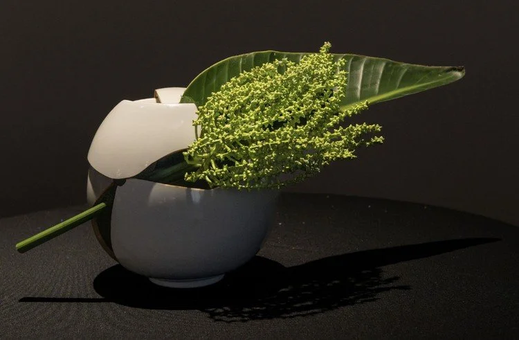 White ceramic vase with a large green leaf and small yellow flowers, casting shadows on a black background.