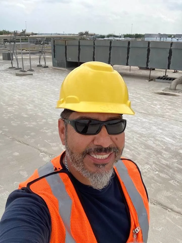 Man wearing yellow safety helmet, sunglasses, and an orange reflective safety vest on a construction rooftop.