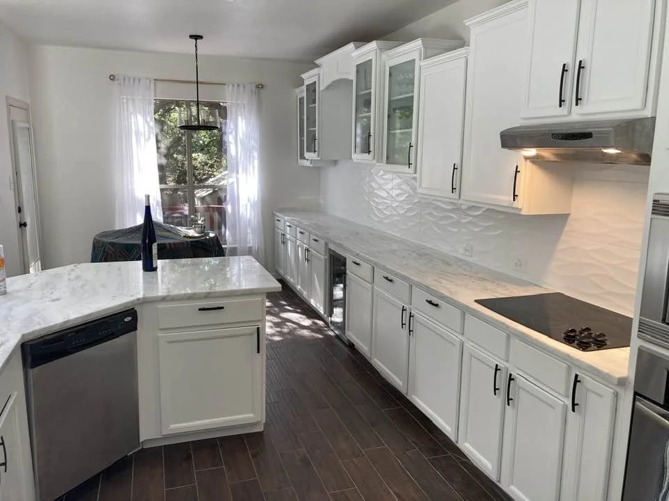 White kitchen with marble countertops, white cabinets with black handles, dark wood flooring, and a dining area with a window and sheer curtains.