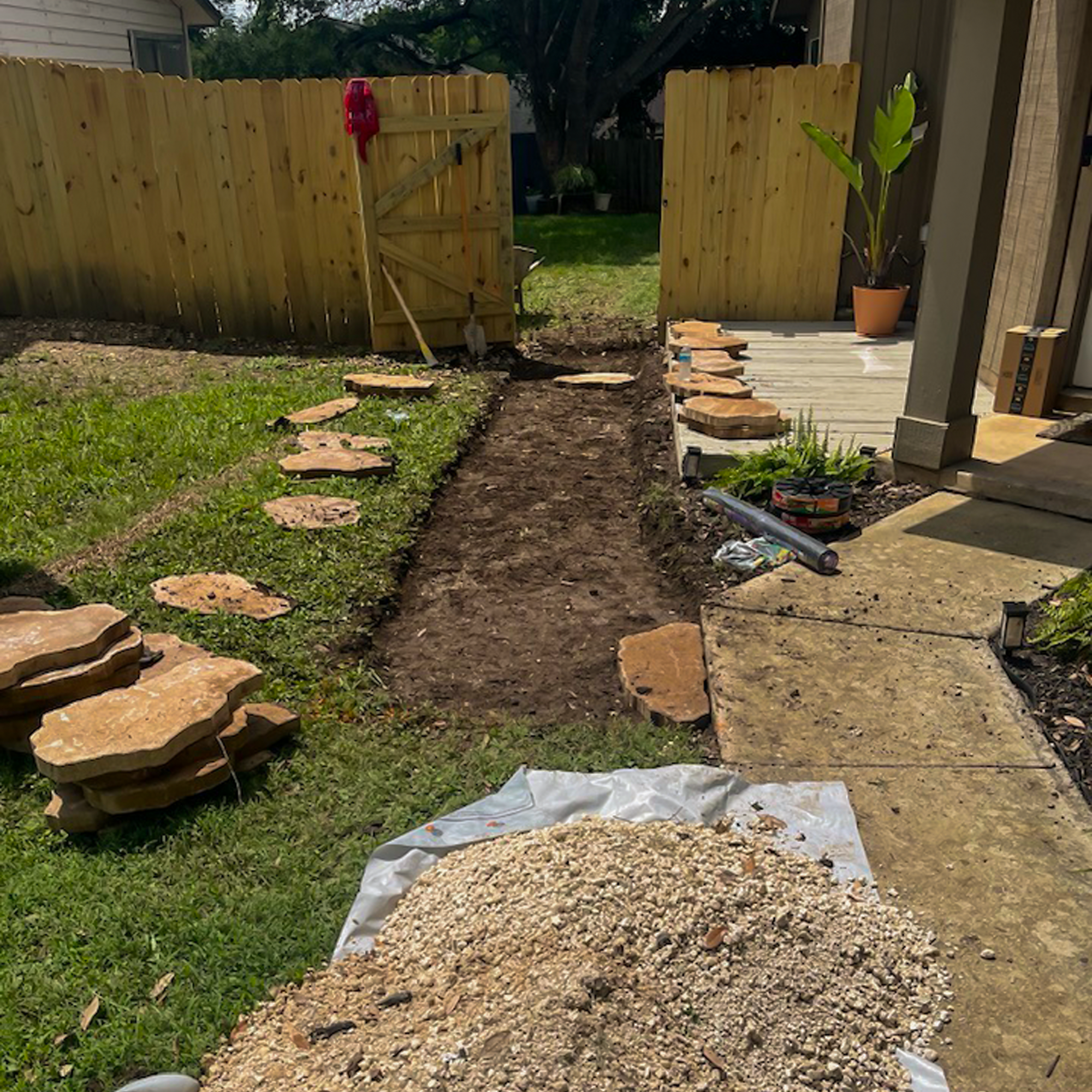 A backyard with a dirt pathway being prepared for a stone walkway, with several large flat stones placed along the edges, a pile of gravel at the front, and a partially finished stone patio on the right side.