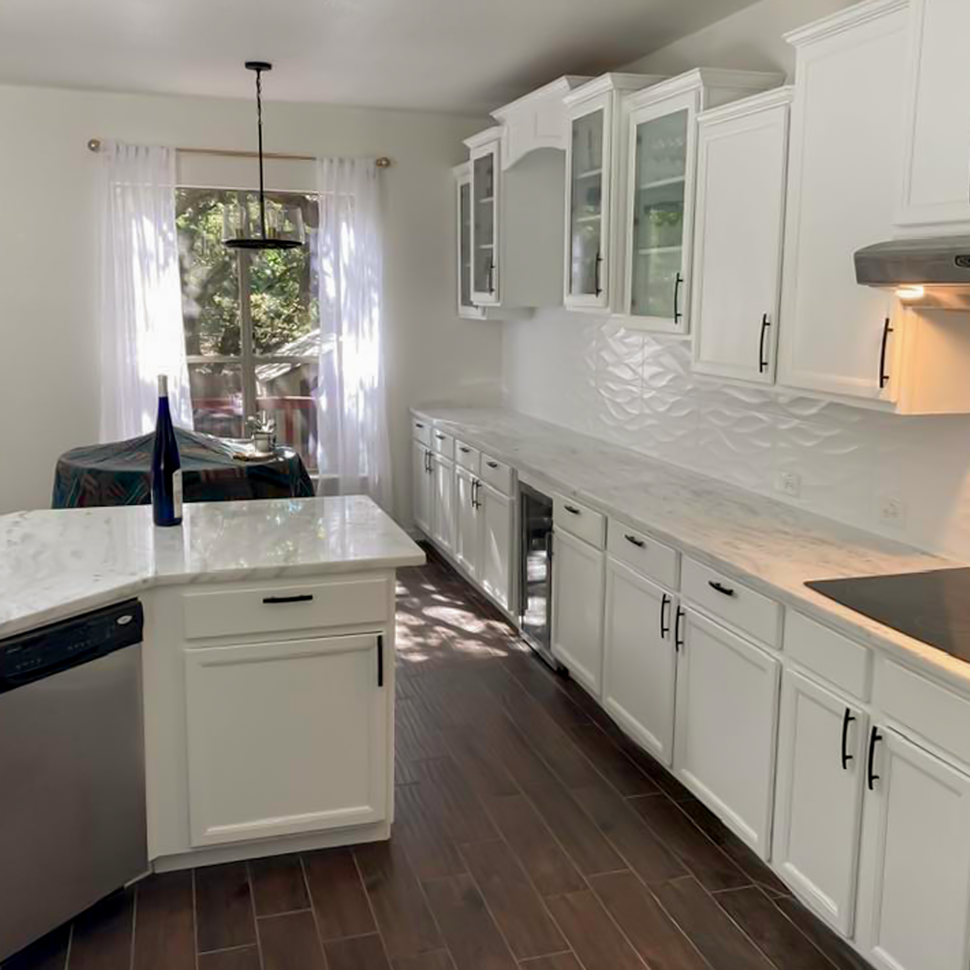 Bright kitchen with white cabinets, marble countertops, and a dark wood floor; a dining table with a blue vase near a large window with white curtains.