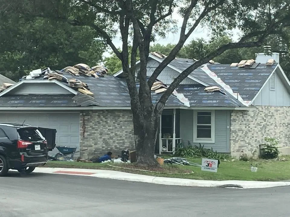 A house under roofing construction with some missing shingles, a large tree in front, and a black SUV parked nearby. There is a sign on the lawn related to construction.