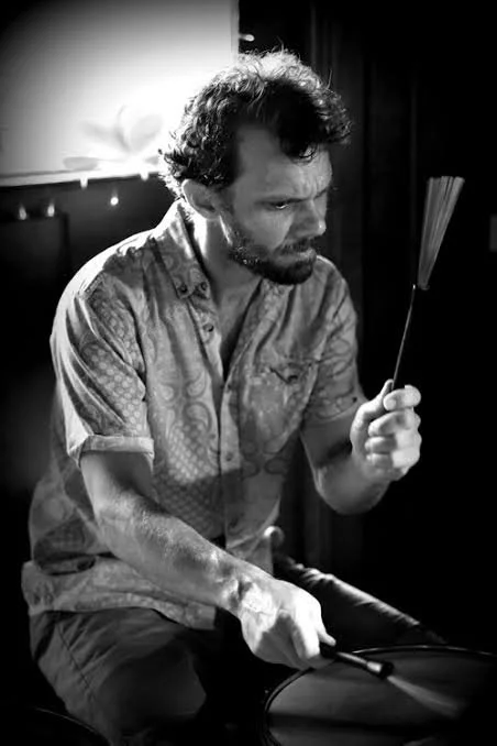 A man with curly hair and a beard, wearing a patterned short sleeve shirt, sitting in a dimly lit room, holding a feather, with a drumstick in his other hand.