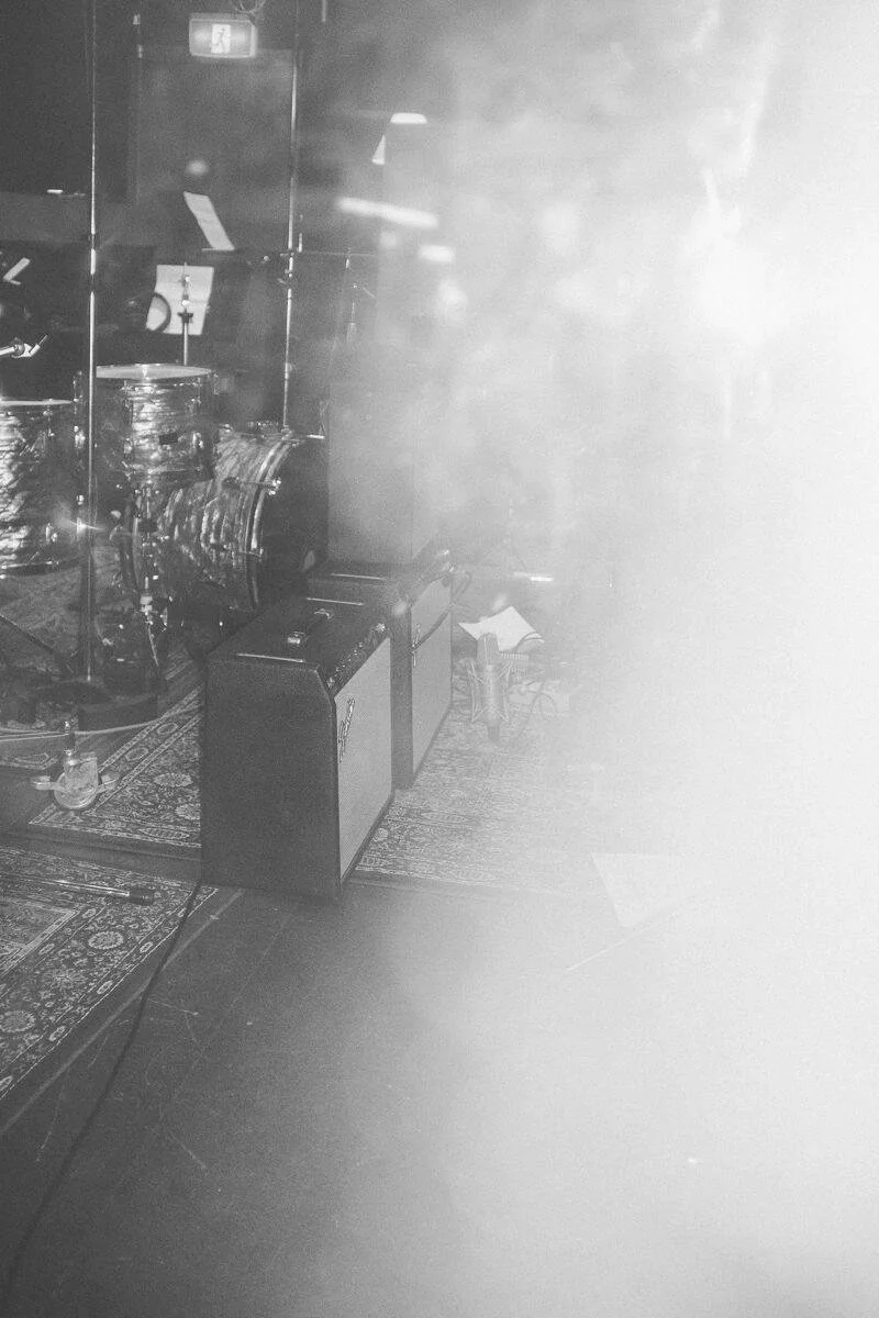 Black and white photo of a music stage with drums, amplifier, and microphone stand, partially obscured by bright light or fog.