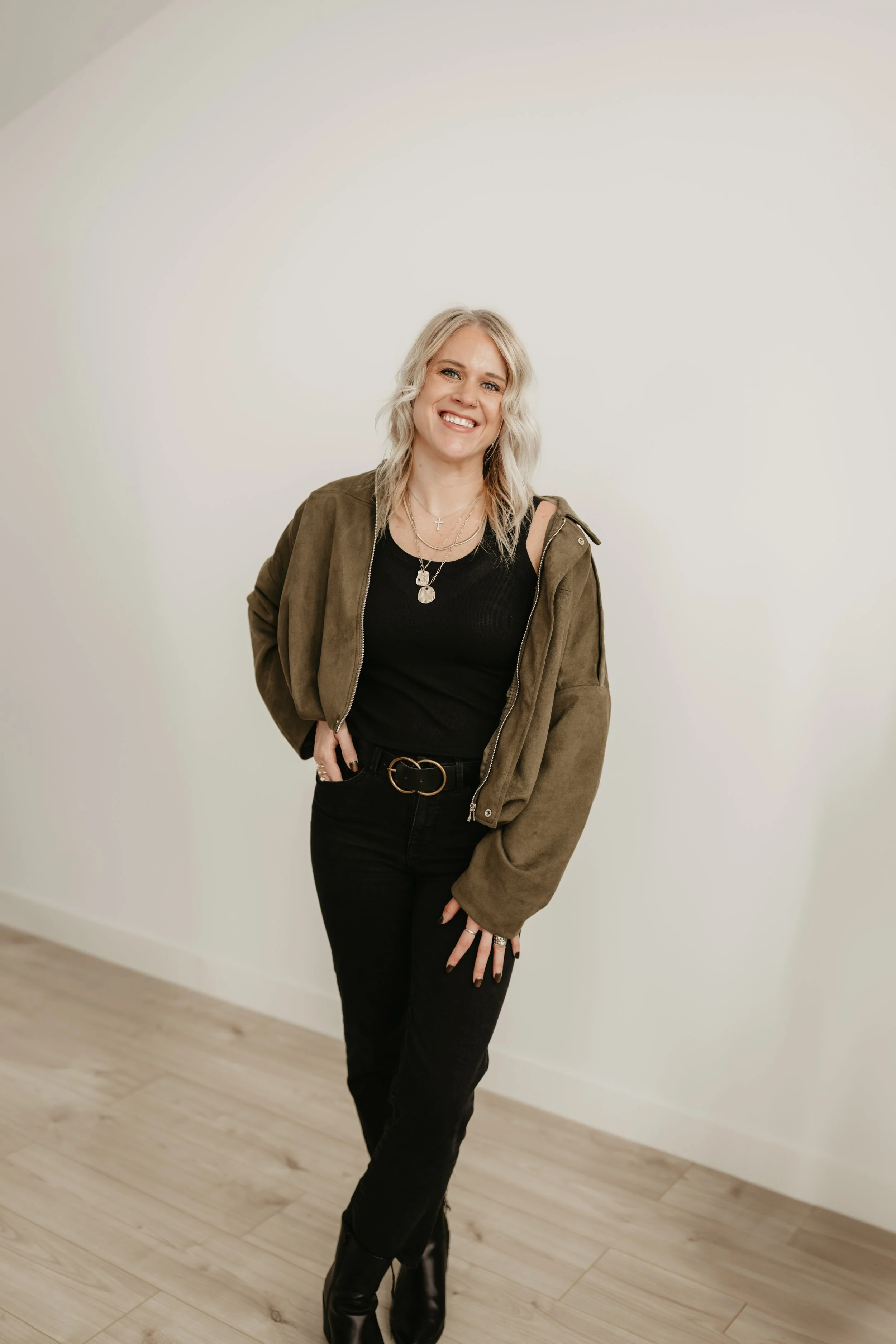 A smiling woman with blonde hair wearing a black top, black pants, a brown jacket, and black boots, standing against a plain white wall.