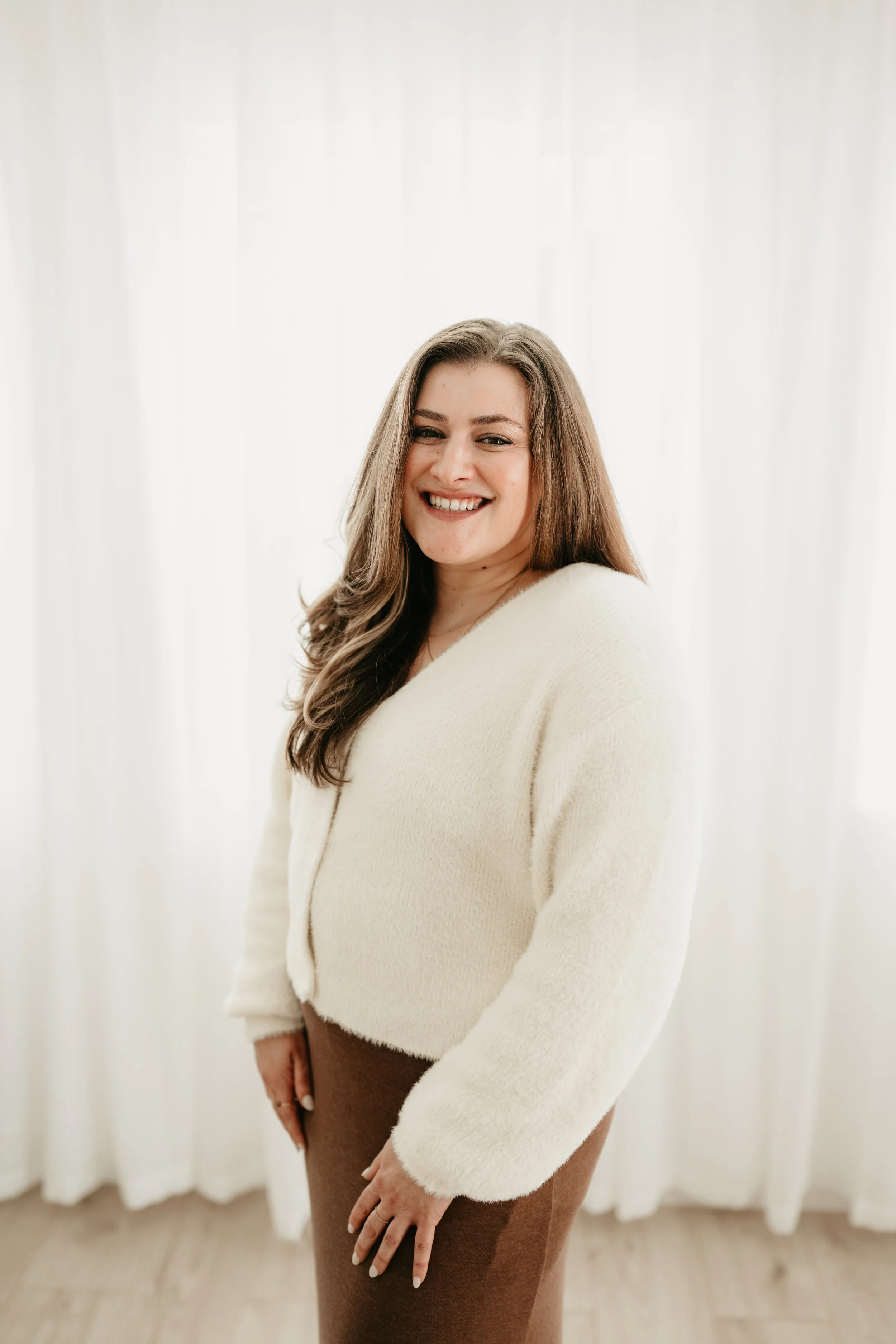 A smiling woman with long brown hair standing indoors with white curtains in the background.