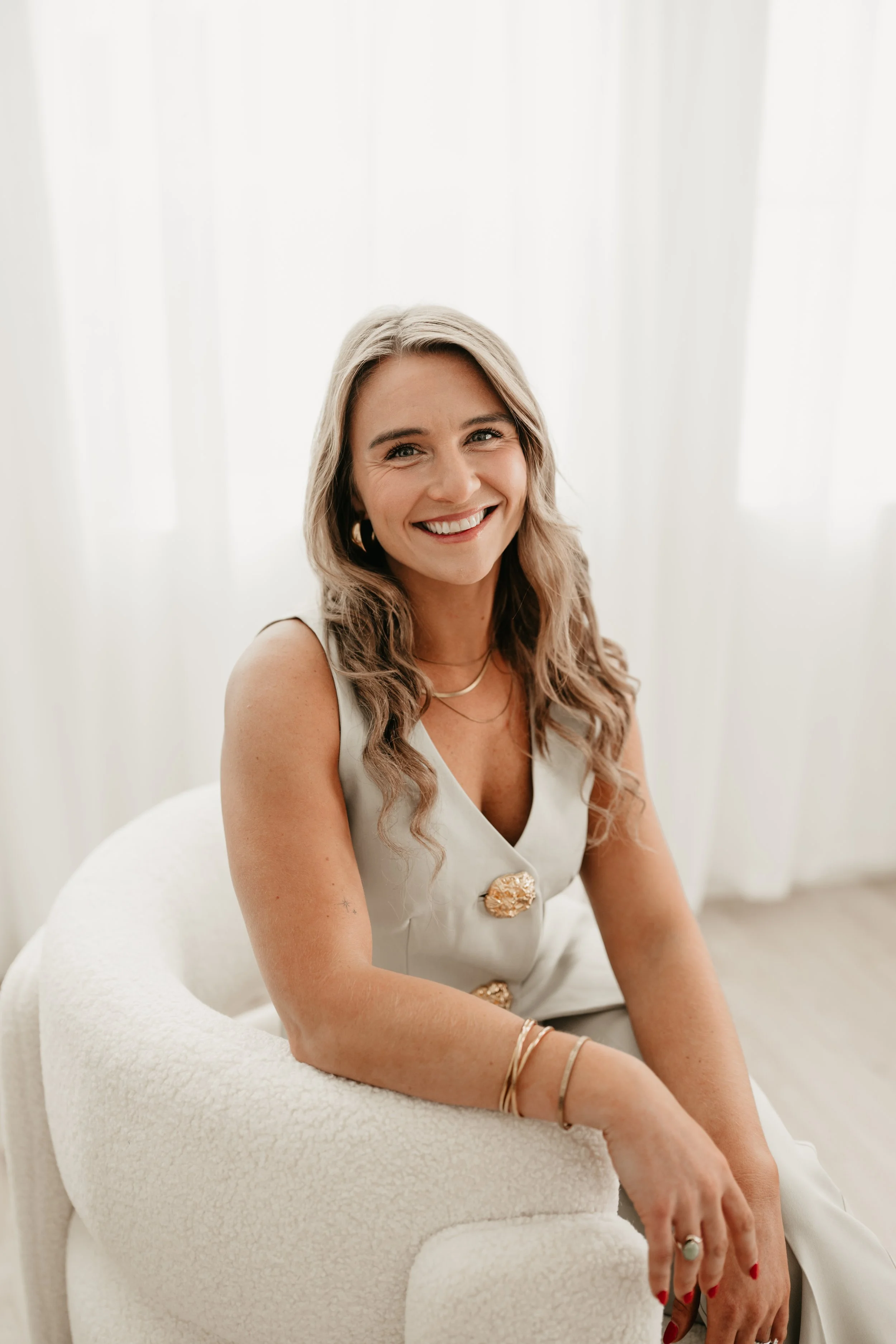 A smiling woman with long, wavy blonde hair wearing a sleeveless white top with large decorative buttons, seated on a white textured chair in front of sheer white curtains.