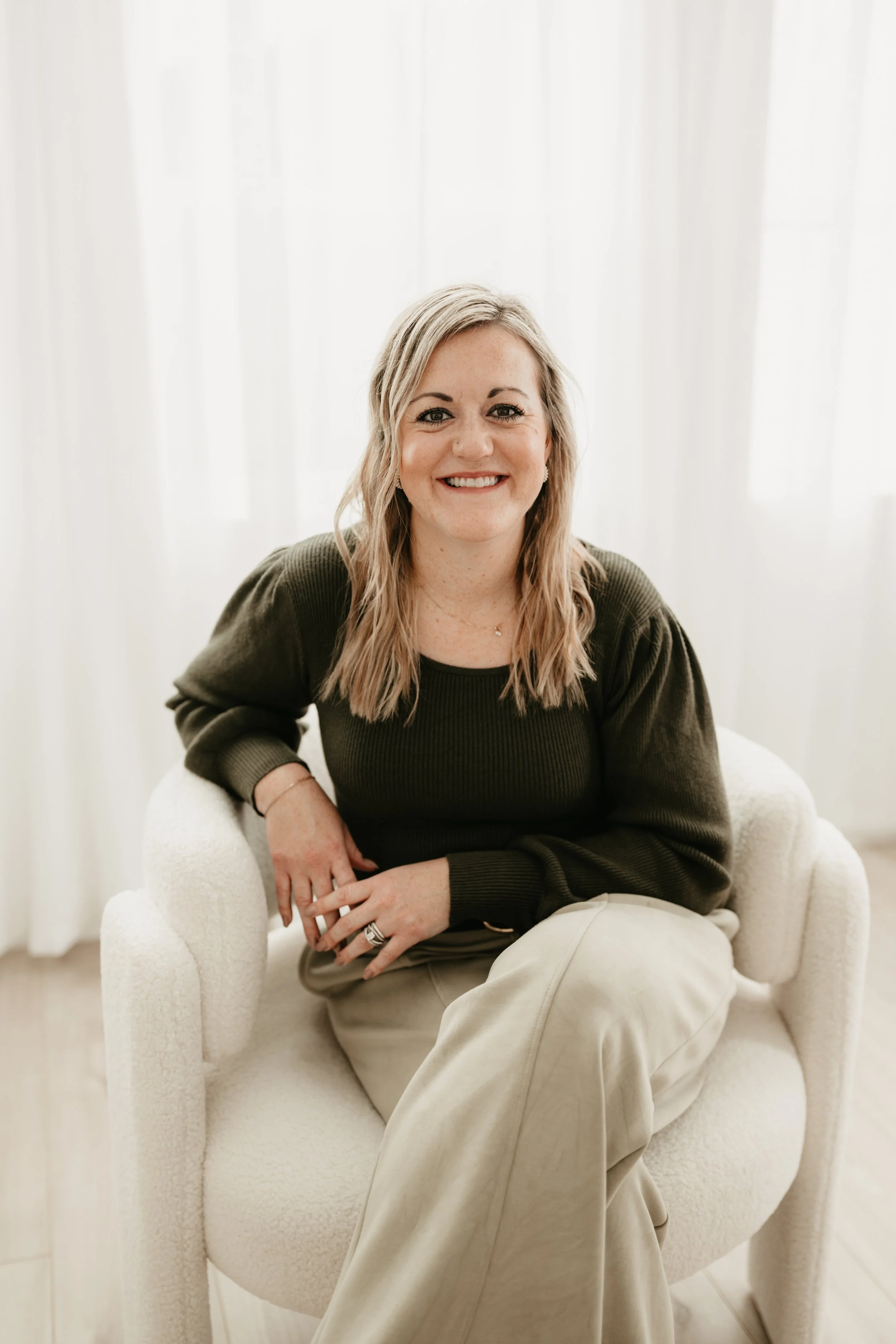 A woman with blonde hair sitting on a white chair, smiling at the camera, in front of a background of white curtains.