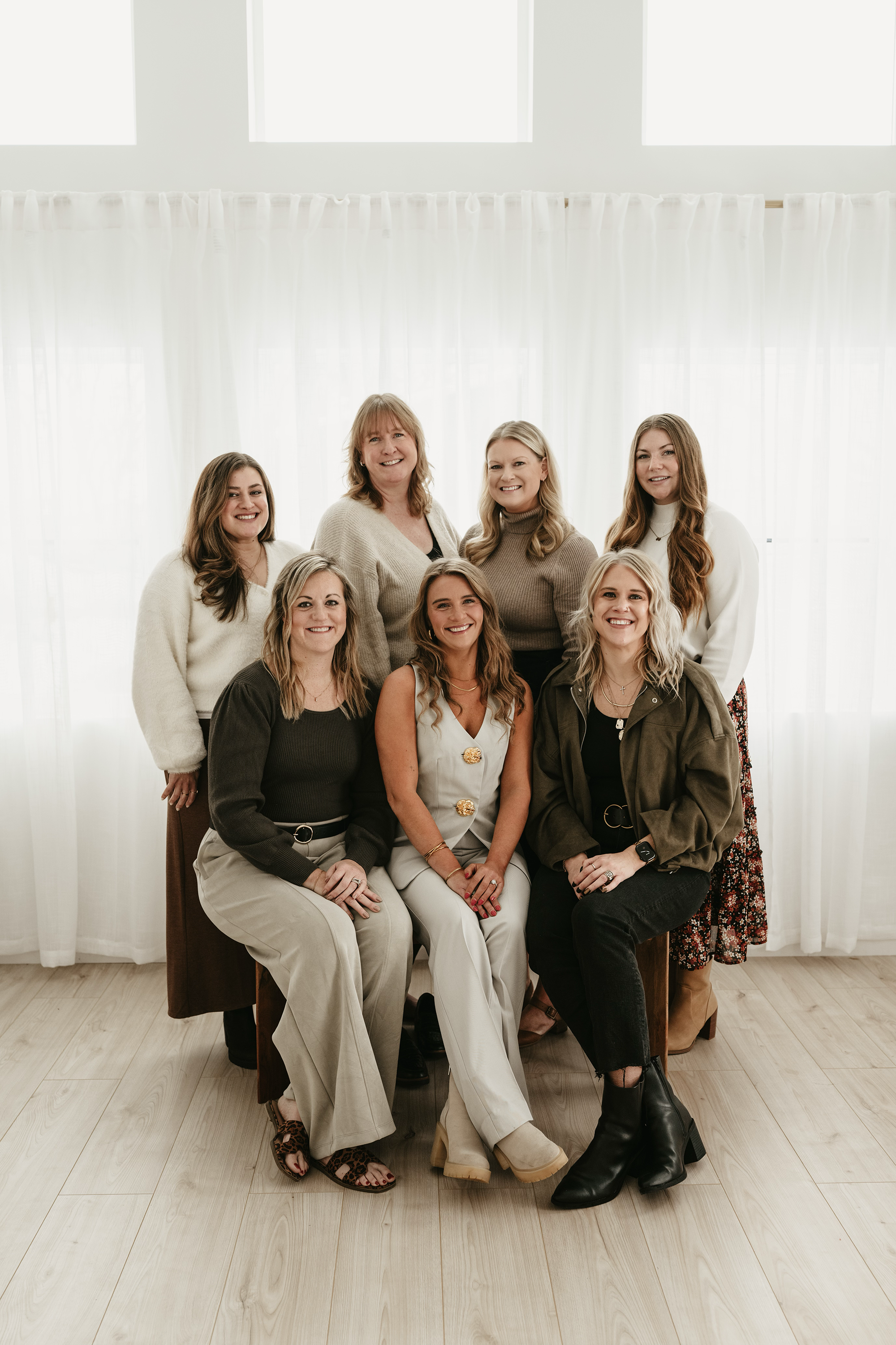 Group of nine women posing together indoors in front of sheer white curtains, smiling at the camera.