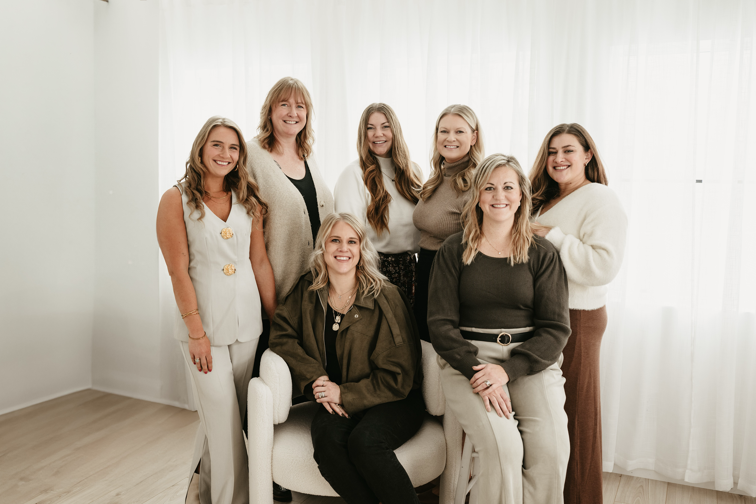 Group of nine women smiling in a room with white curtains, some sitting and some standing.