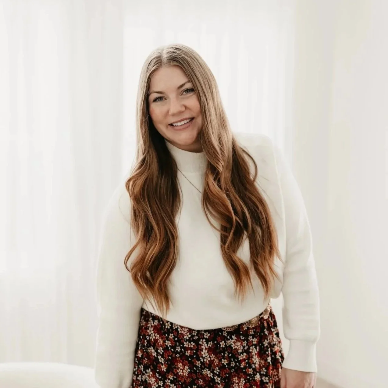 A woman with long, wavy brown hair wearing a white sweater and a floral skirt, smiling in front of a light background.
