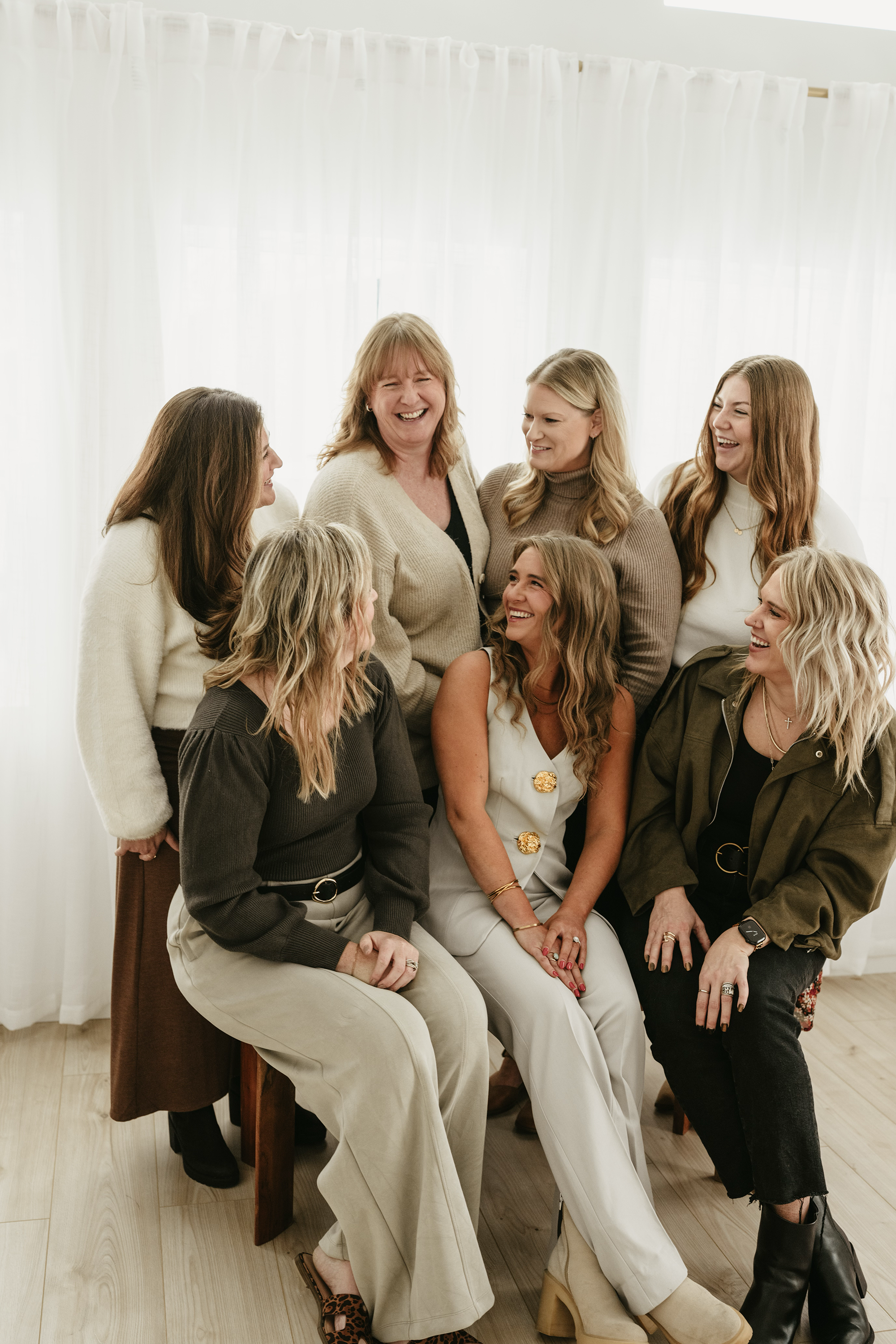 Group of women smiling and laughing together indoors with white curtains in the background.