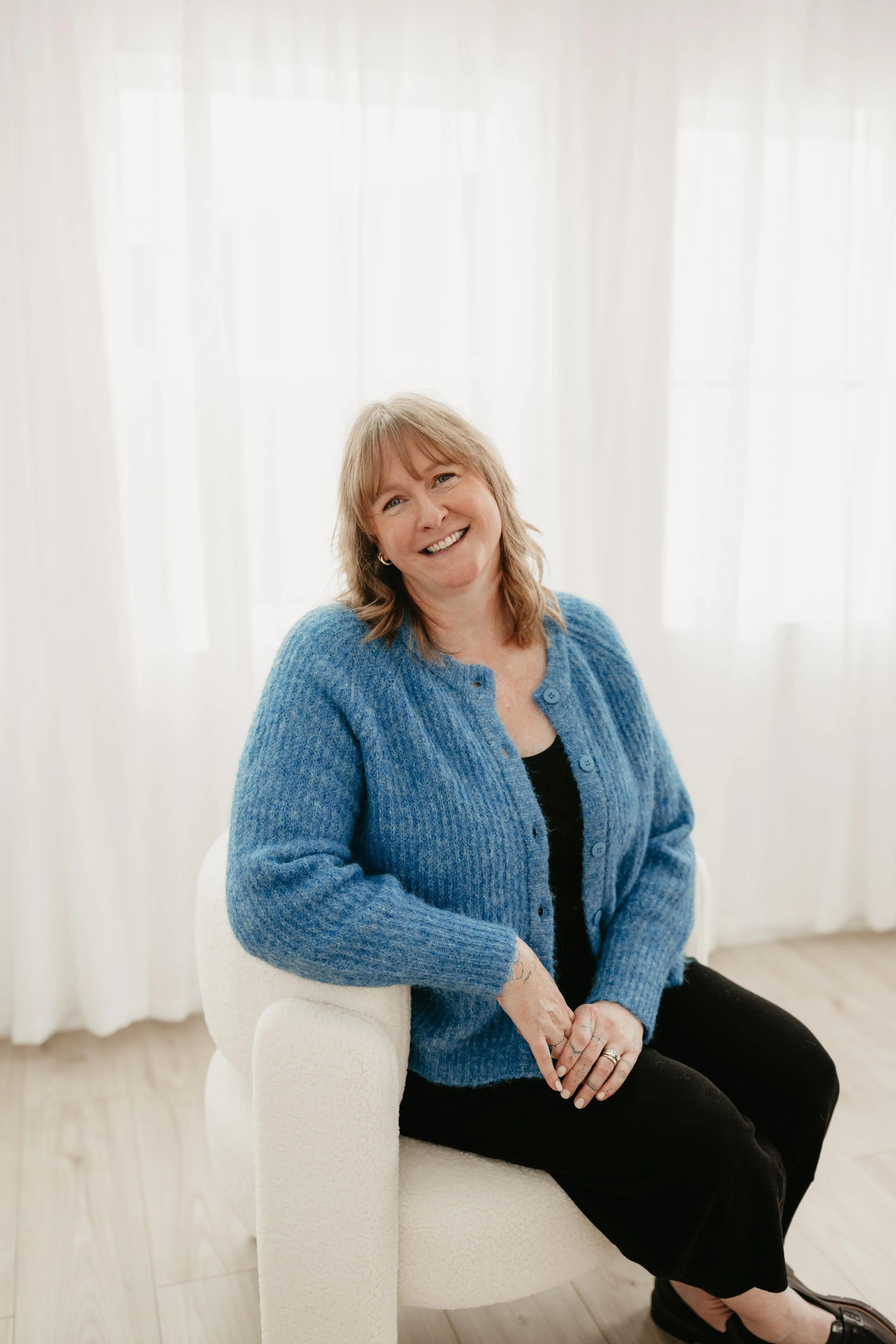 A woman with shoulder-length light brown hair, wearing a blue knit cardigan and black pants, sitting in a white chair in front of sheer white curtains and smiling at the camera.