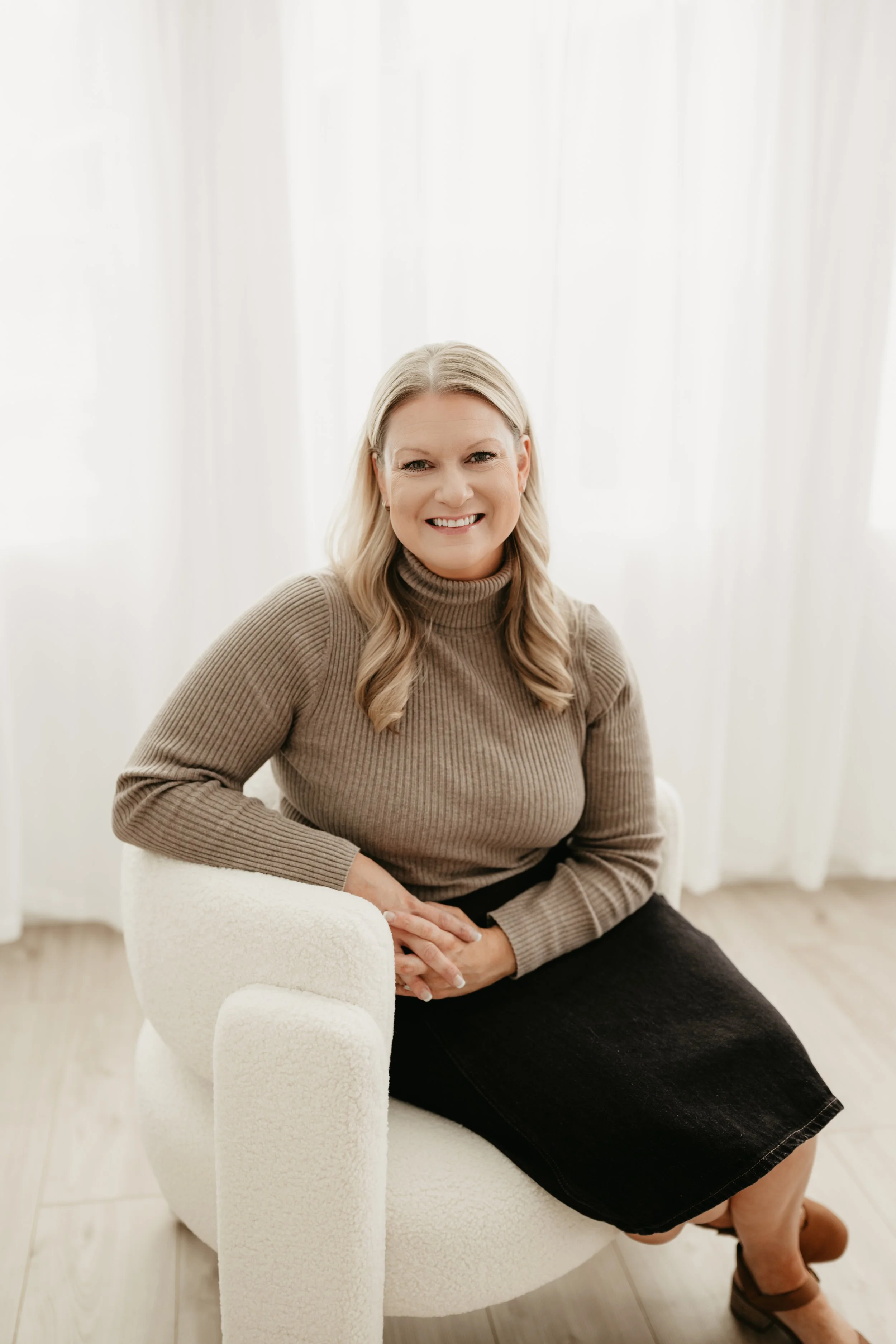 A woman with blonde hair sitting on a white, textured armchair in a bright room with white curtains, wearing a brown turtleneck sweater and a black skirt, smiling at the camera.