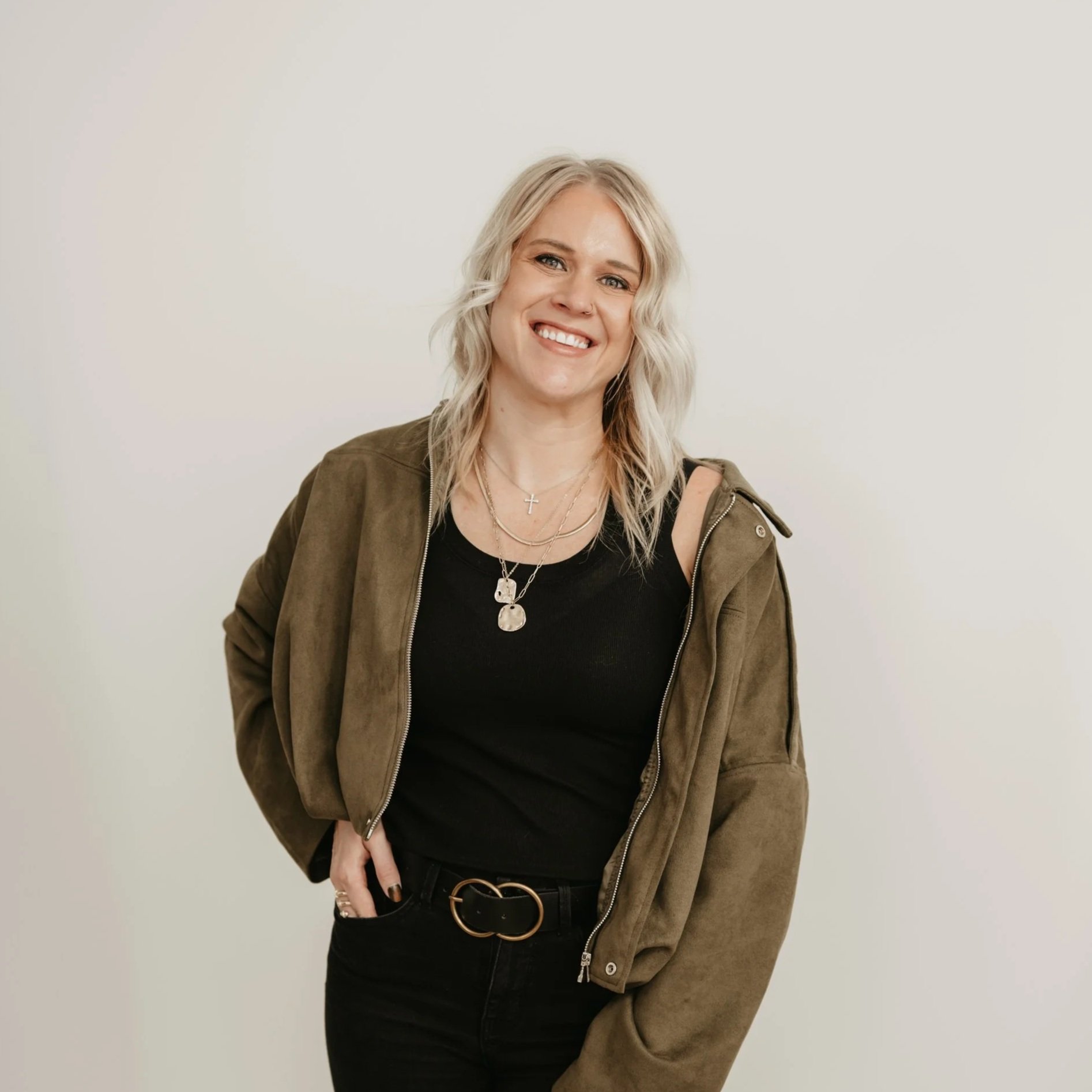 A smiling woman with blonde hair in a casual outfit standing against a plain light-colored background.