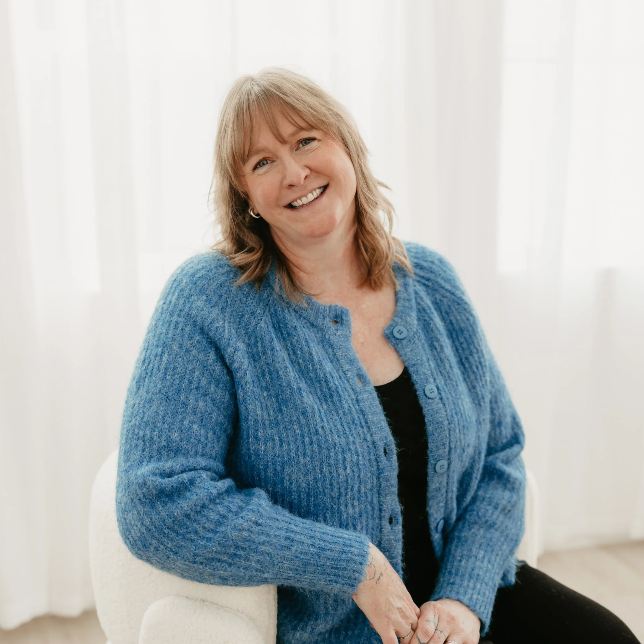 A woman with light brown hair smiling while sitting on a white chair in front of white curtains, wearing a blue cardigan and black top.