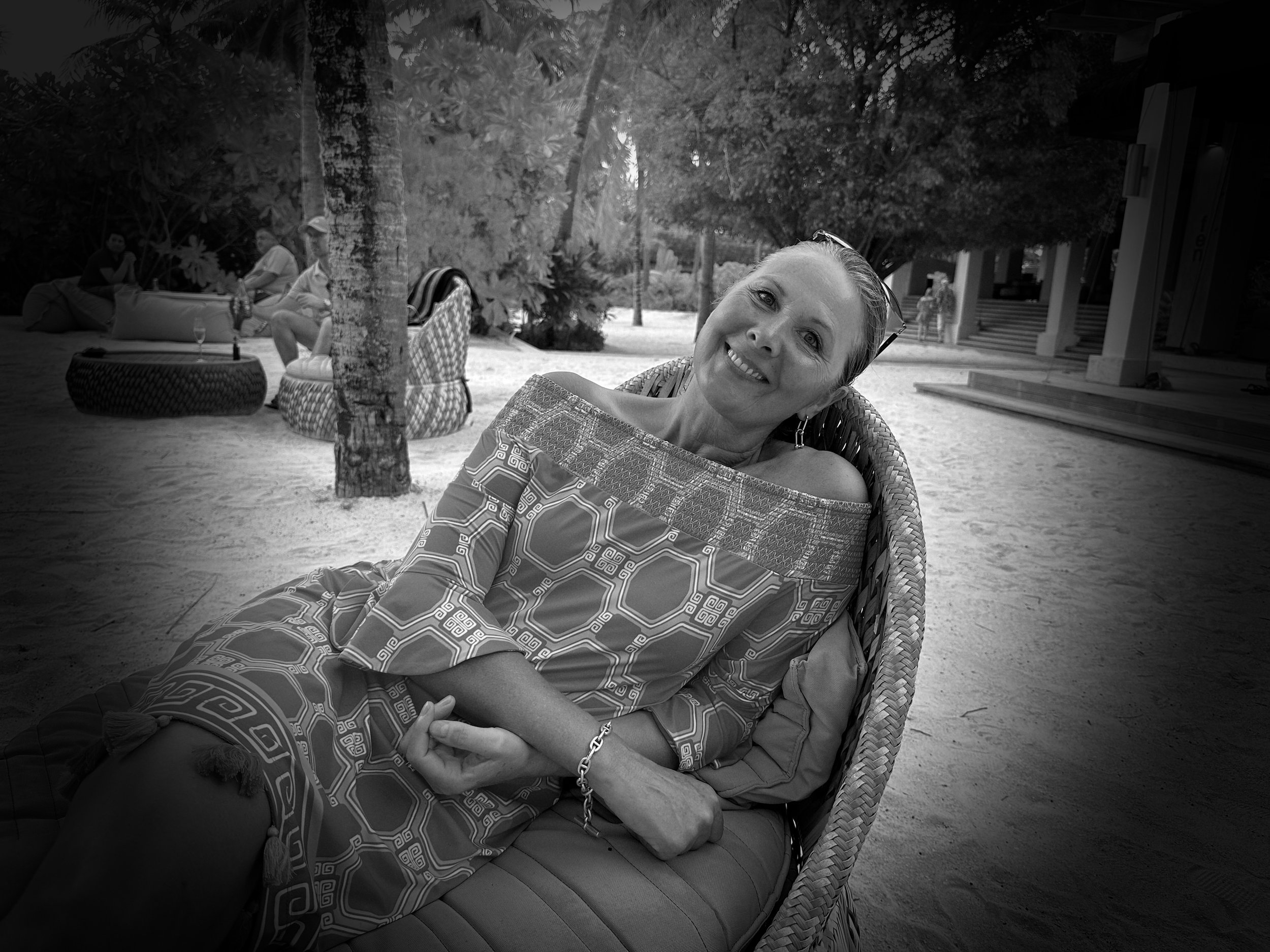 A woman with sunglasses on her head smiling and relaxing on a wicker chair outdoors on a sandy ground, with trees and other people in the background.