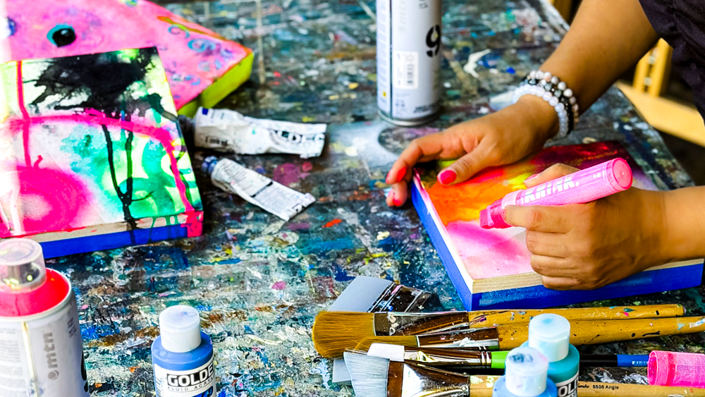 Artist working on a colorful abstract painting with brushes, markers, and paint supplies on a messy cluttered table.