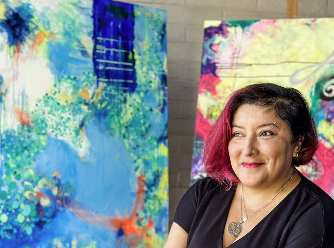 A woman with pink and black hair, wearing a black top, sitting in front of colorful abstract paintings.