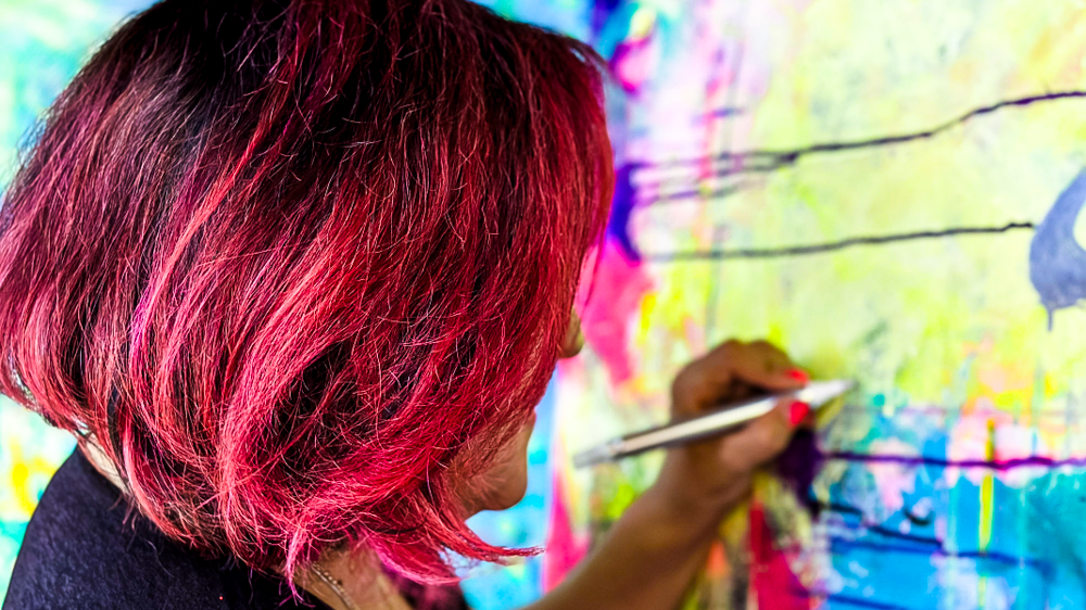 Woman with short hair painted in shades of pink and red, painting on a colorful abstract mural with a brush.