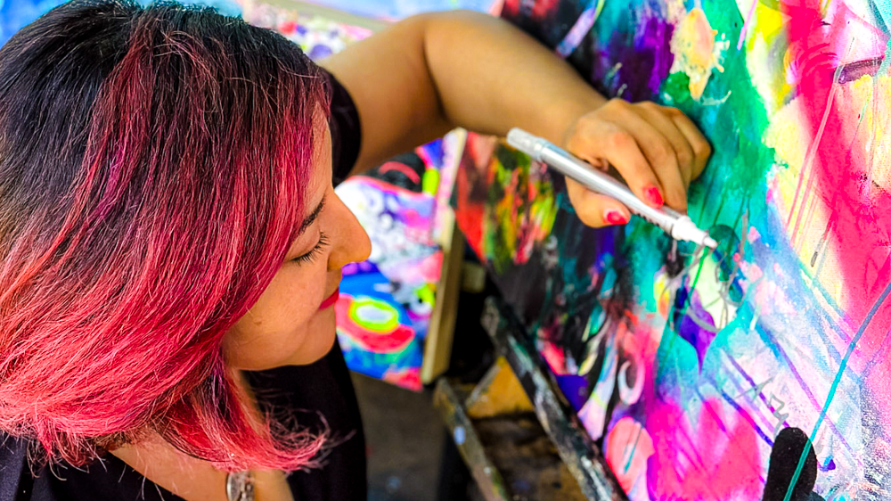 A woman with pink and black hair painting abstract colorful artwork on a large canvas.