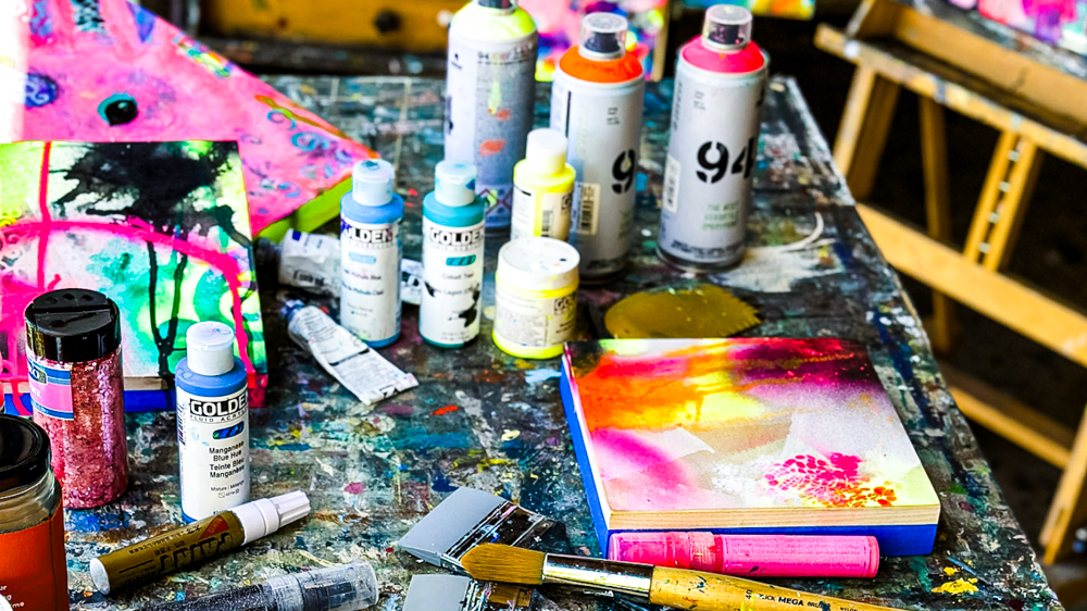 Assorted bottles of acrylic paint, a paintbrush, a small abstract canvas, and a pink and yellow painted notebook on a cluttered artist's worktable.