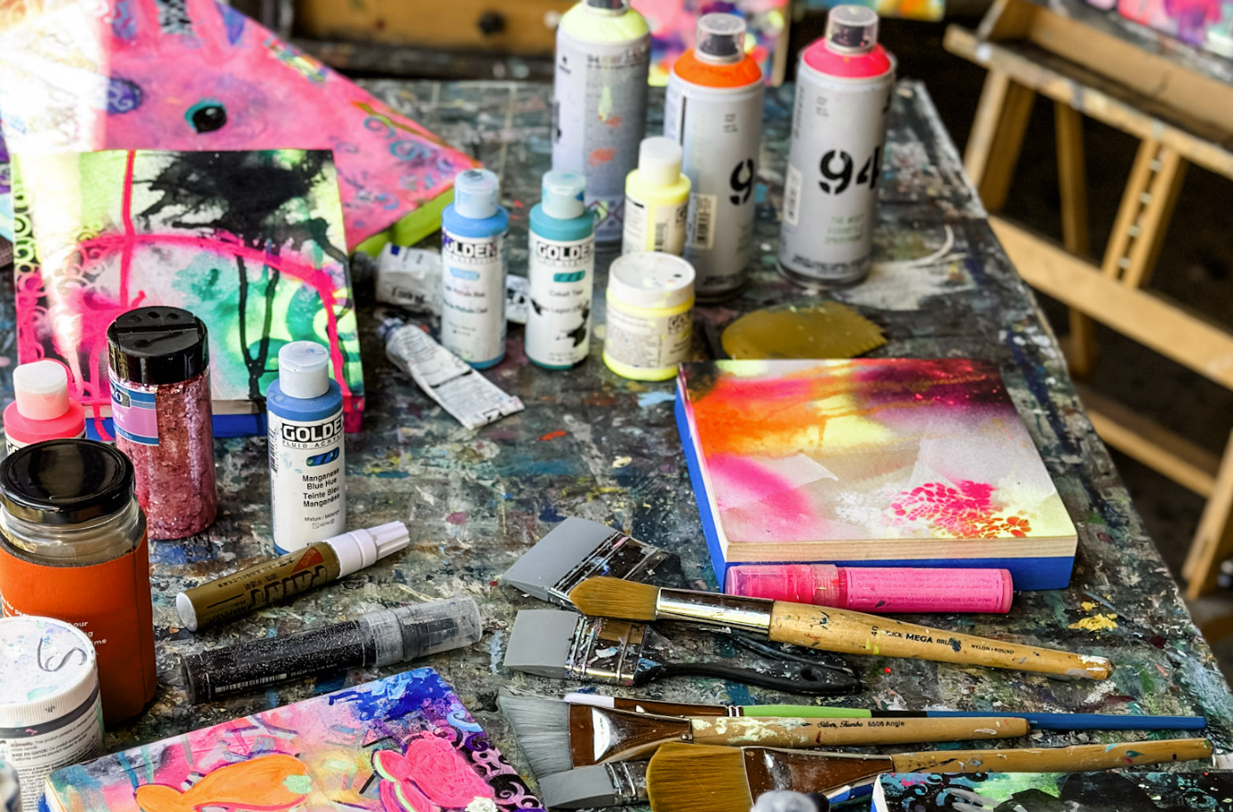 An art workspace covered with various acrylic paint bottles, brushes, and colorful abstract paintings on small canvases.