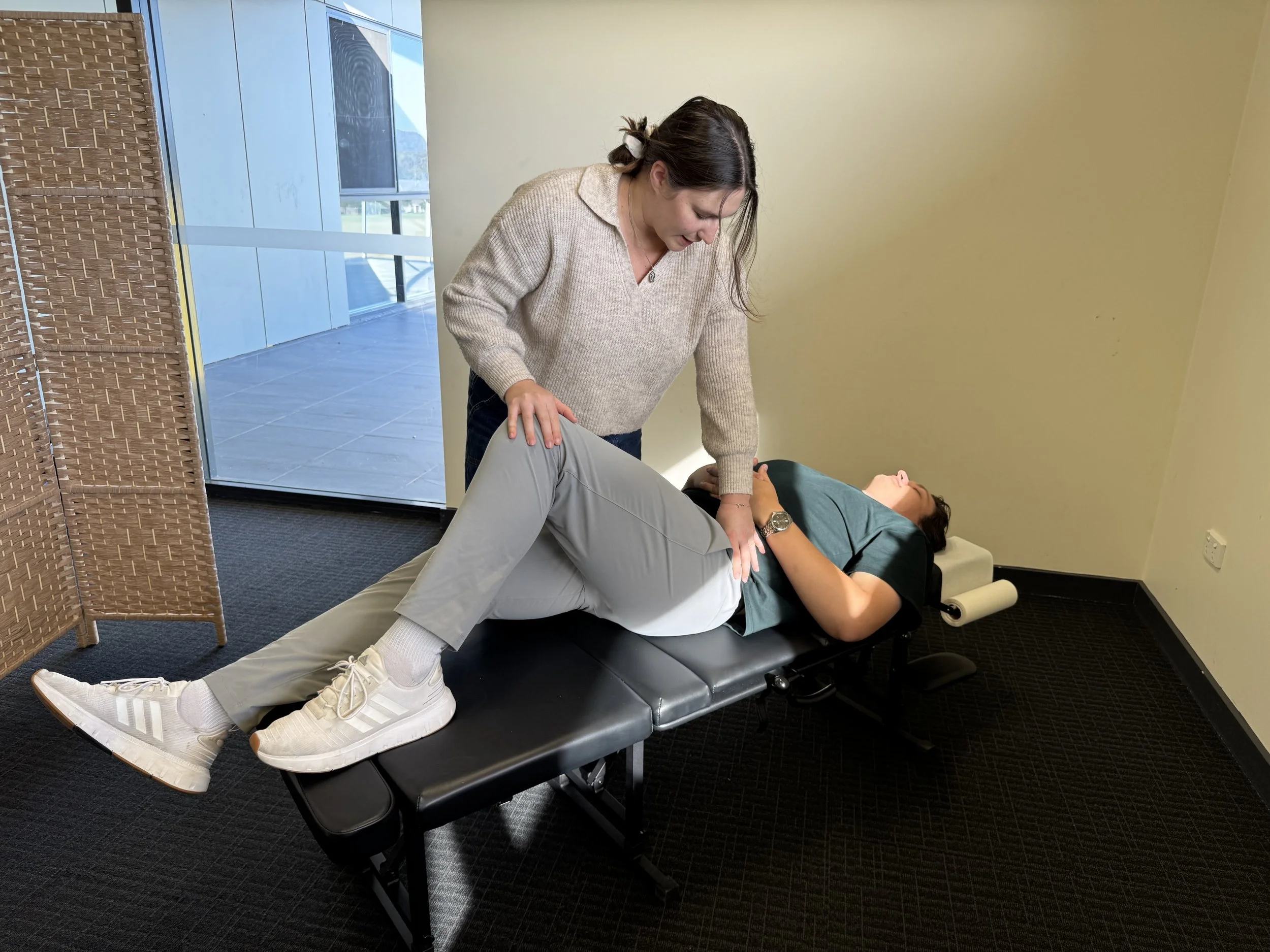 A woman in casual clothes performing a physical therapy or chiropractic adjustment on a man lying on a treatment table in a clinic room with a large window and a decorative divider.