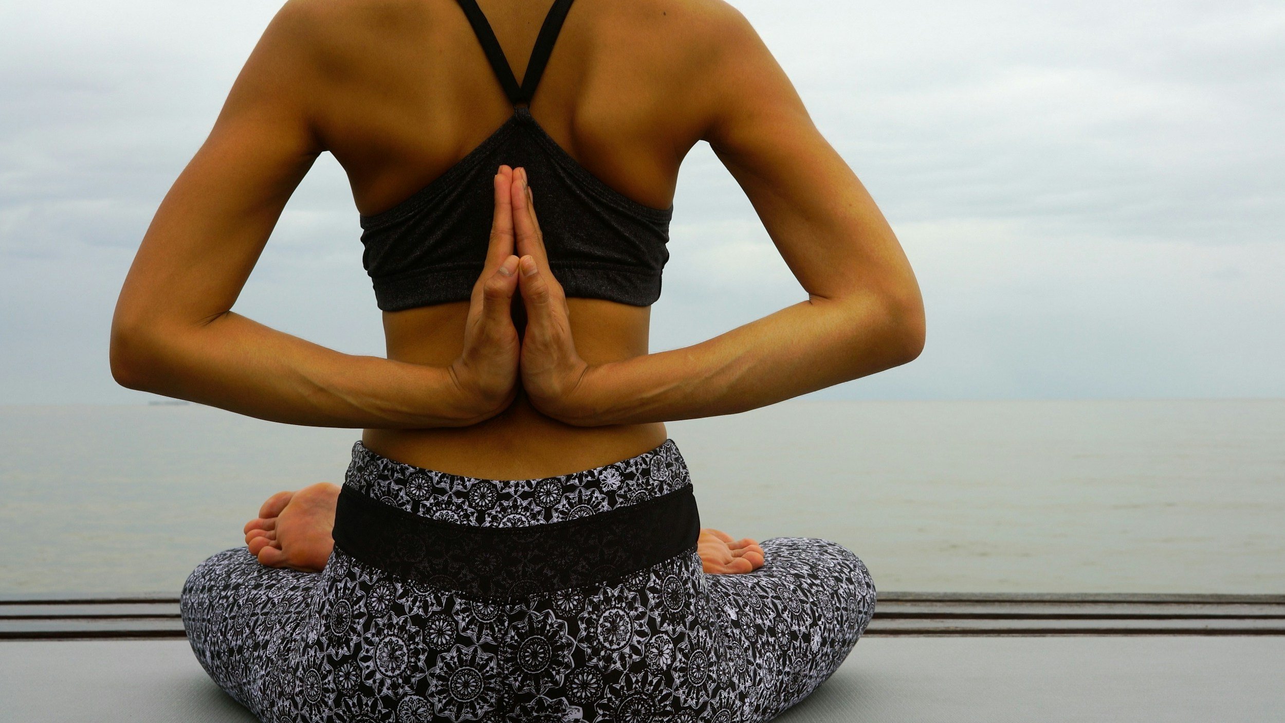 A person practicing yoga outdoors by the water, sitting in a cross-legged position with hands in a prayer pose at the chest, facing away from the camera, in front of a cloudy sky.