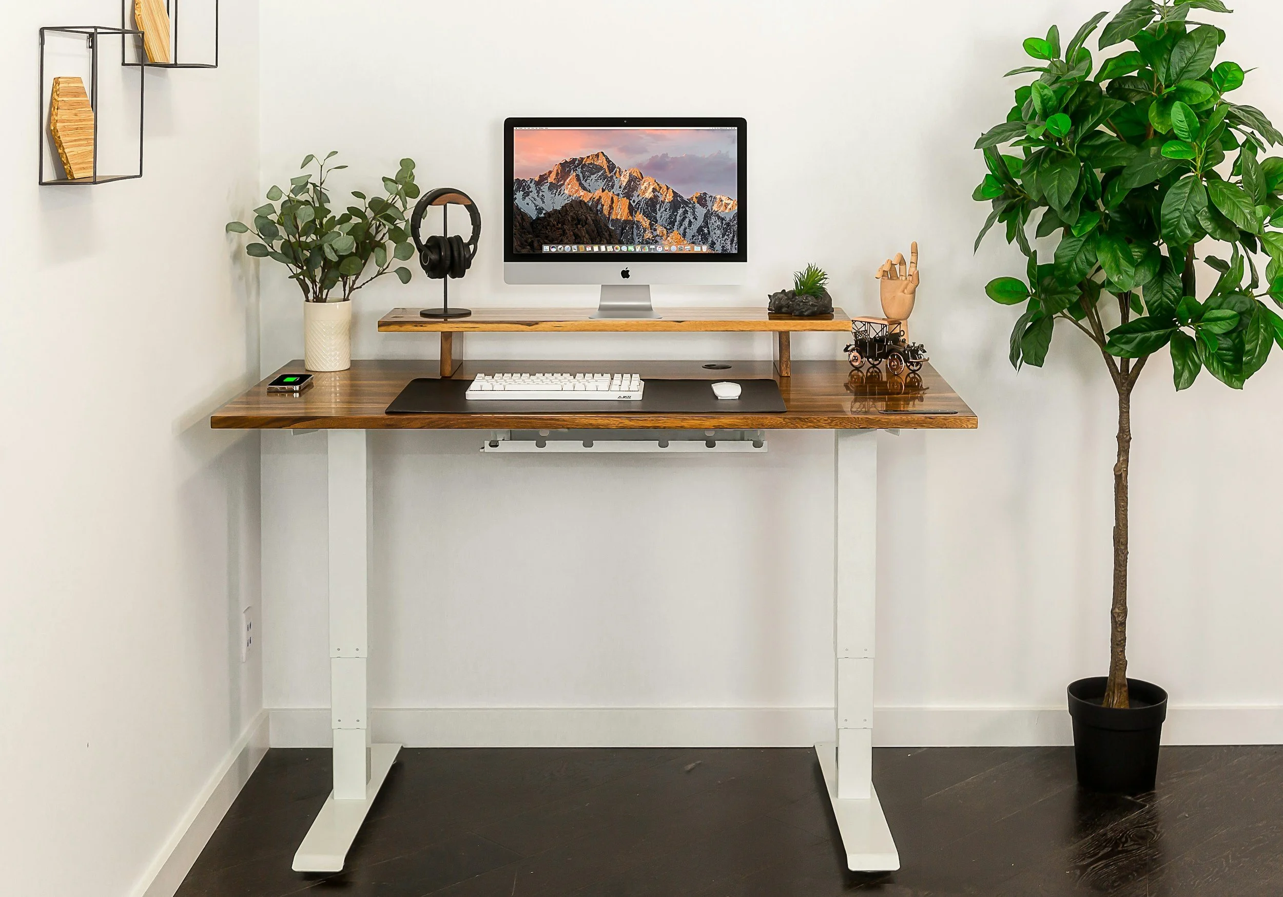 A home office setup with a wooden desk on white metal legs, a computer monitor, a keyboard, a mouse, a pair of headphones, and various decorative items including potted plants, a miniature stroller, and wall shelves with books, all against a white wall with dark flooring.