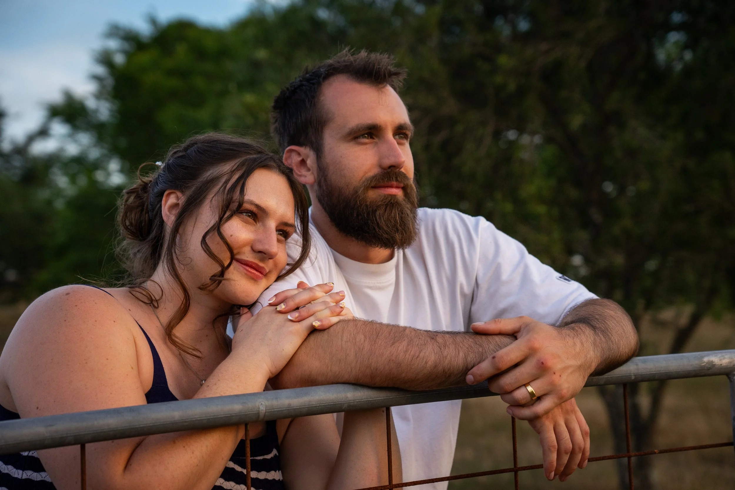 A young woman and a man standing behind a metal fence, looking into the distance, with trees in the background.