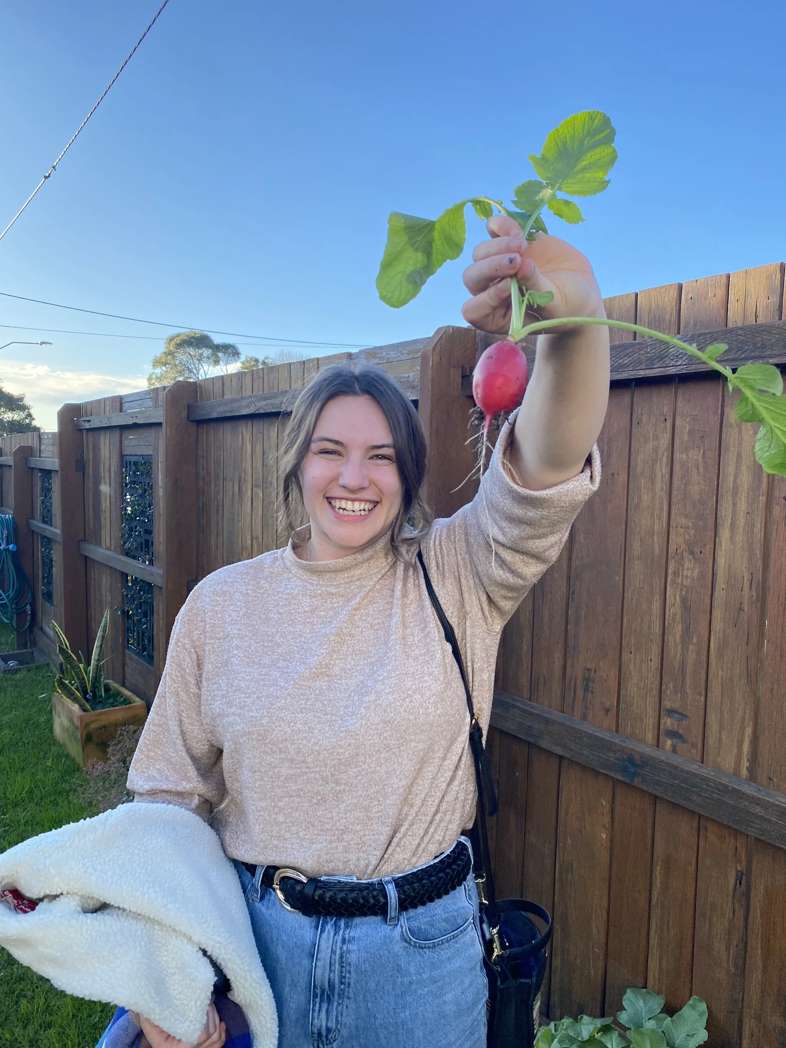 A woman smiling outdoors holds up a freshly picked radish connected to its leafy green top. She is standing in front of a wooden fence on a sunny day.